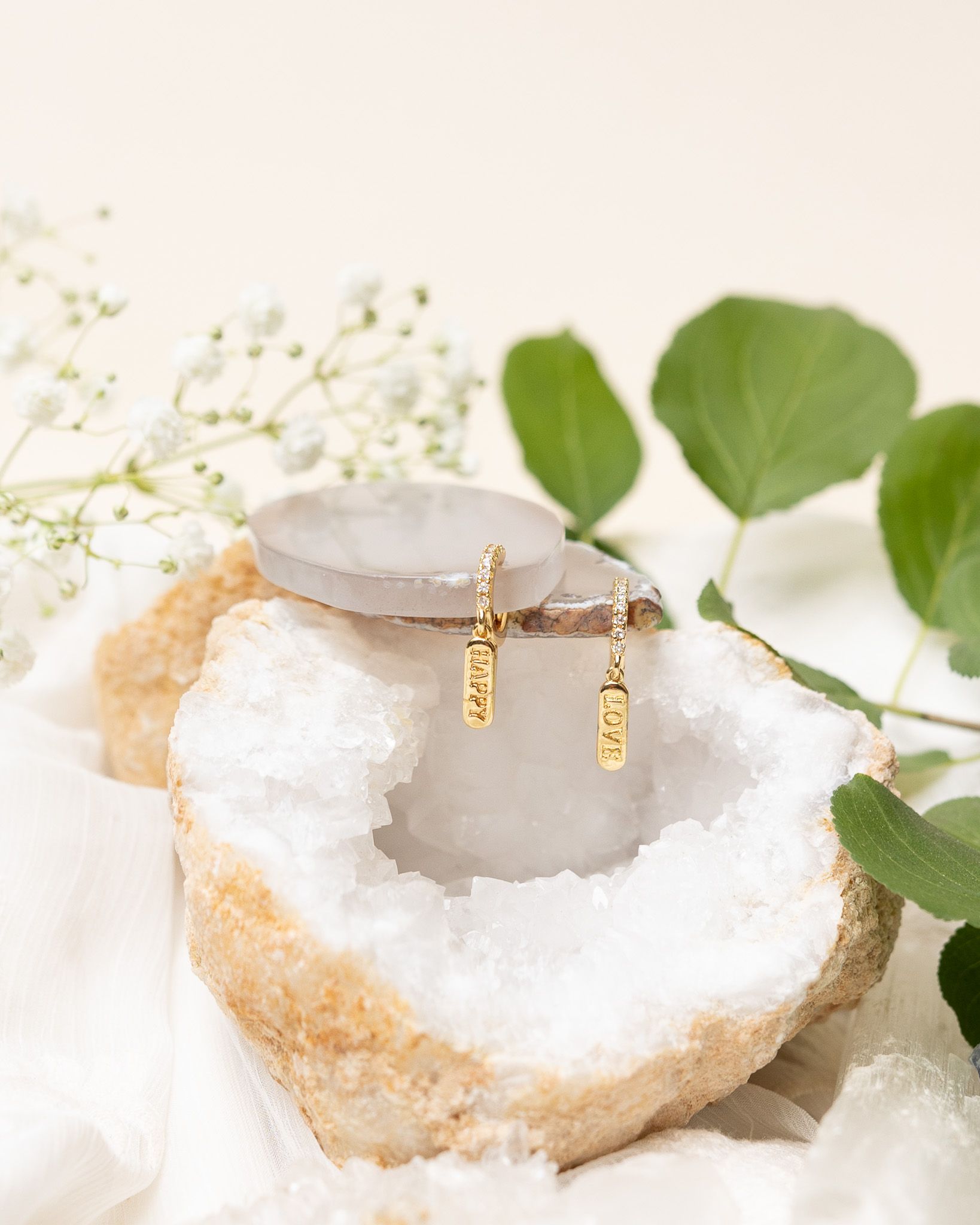 Gold charm earrings displayed on white crystal geode with green leaves and baby's breath.