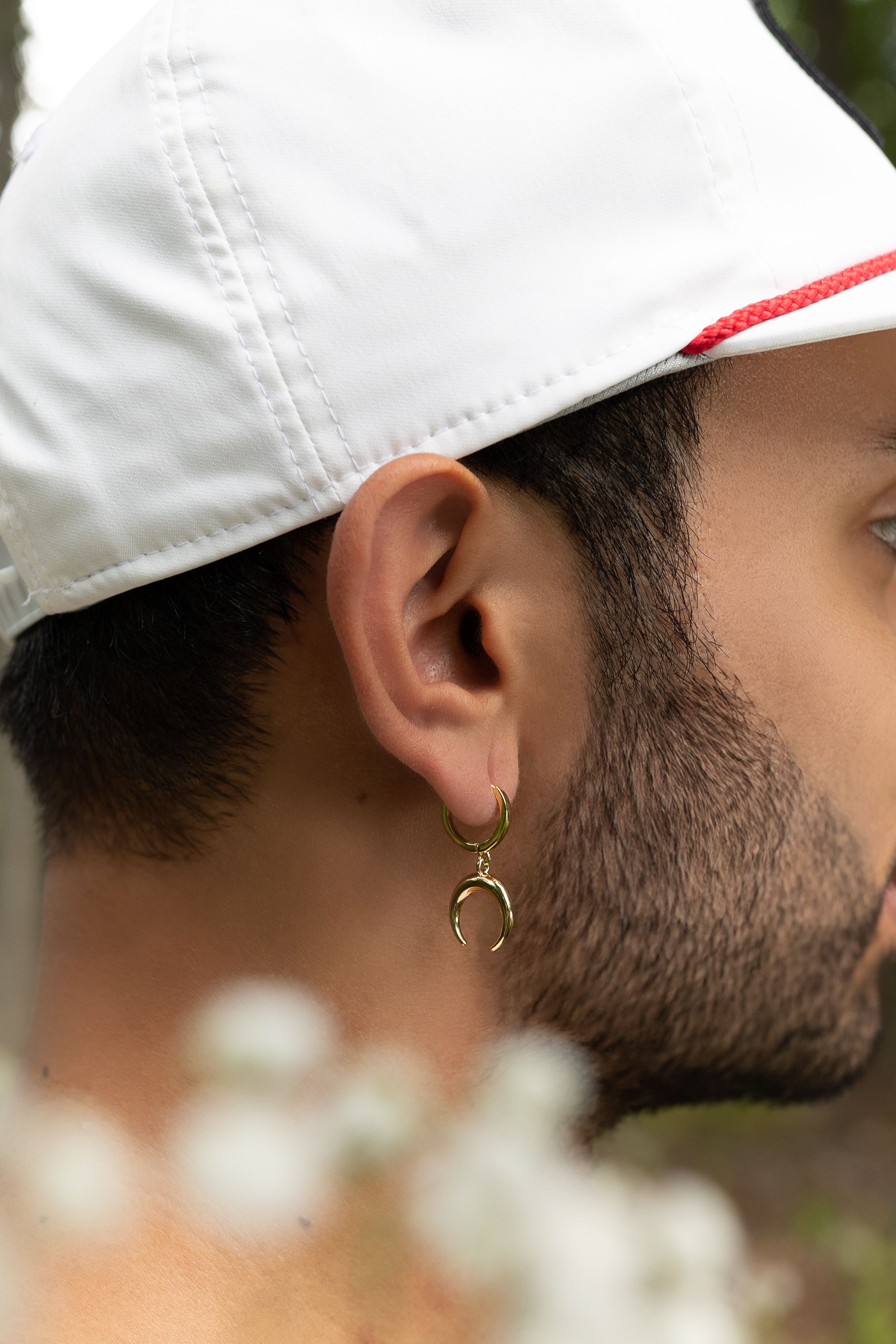 Close-up view of a person wearing white cap and Stay Gold Collective crescent moon earring against blurred floral background.
