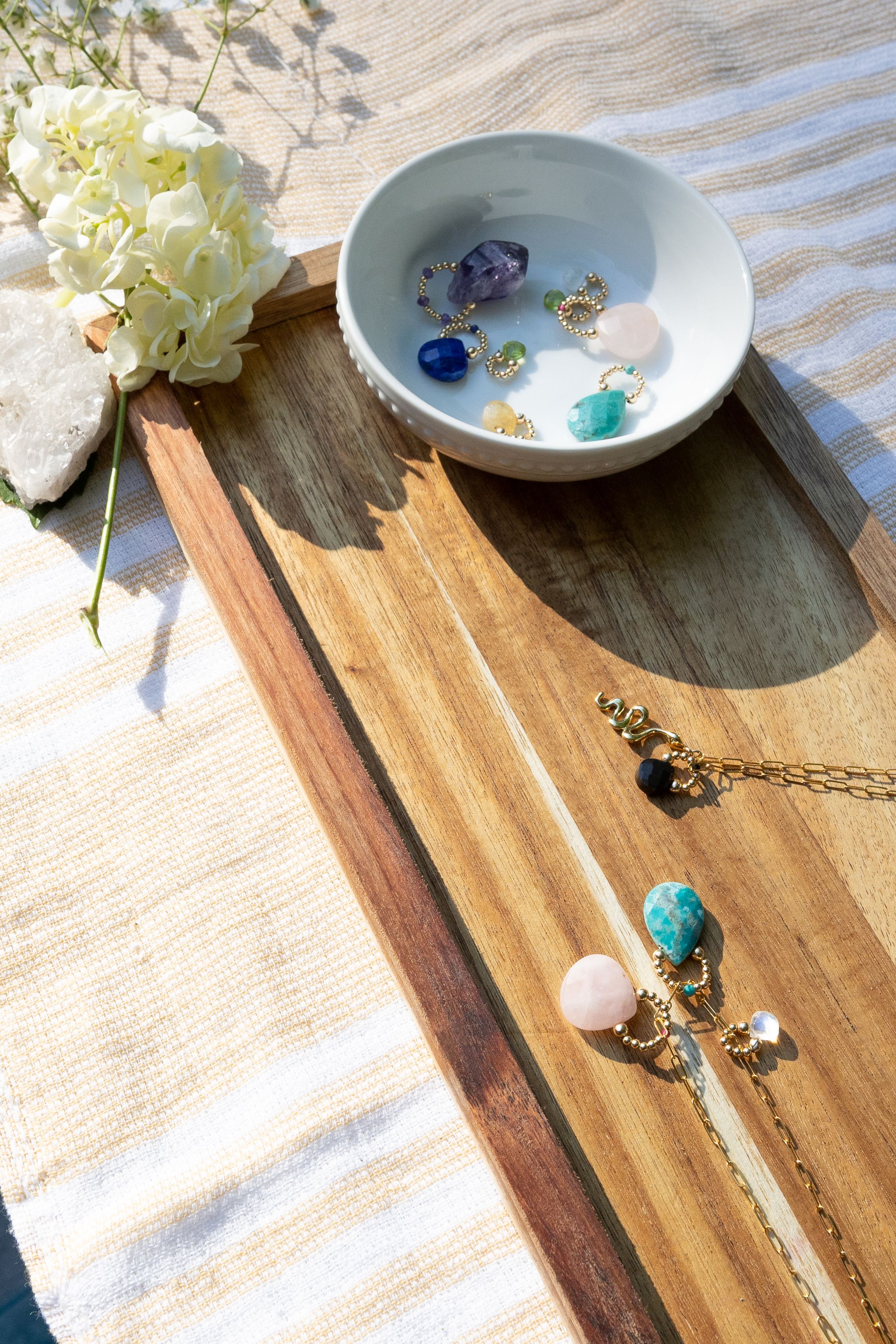 Jewelry display with small ceramic bowl and wooden board featuring gemstone accessories and white flowers.