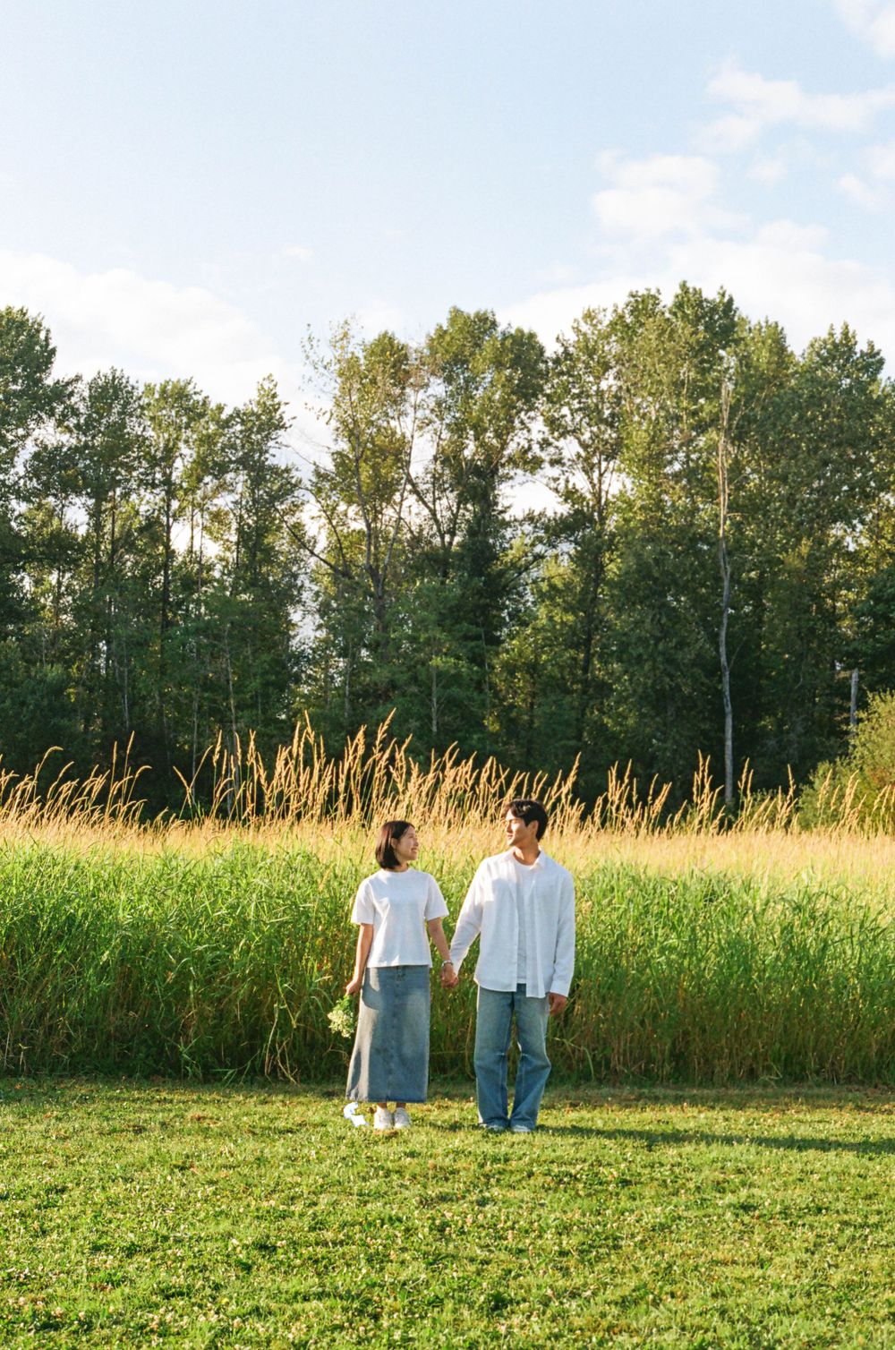 Two people walking hand in hand through a grassy field with trees in the background.