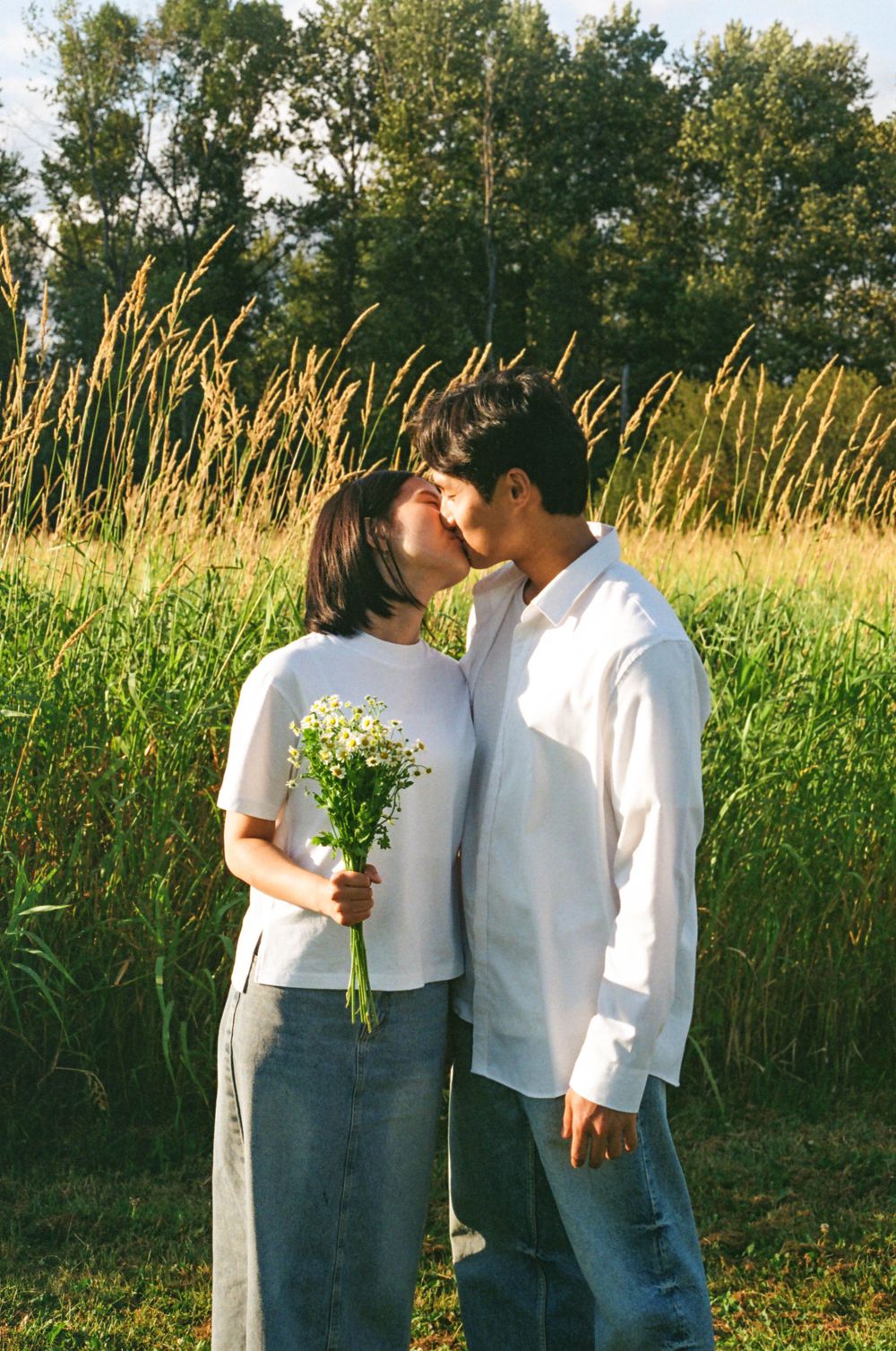 A couple shares an intimate moment while holding wildflowers in a sunny field.
