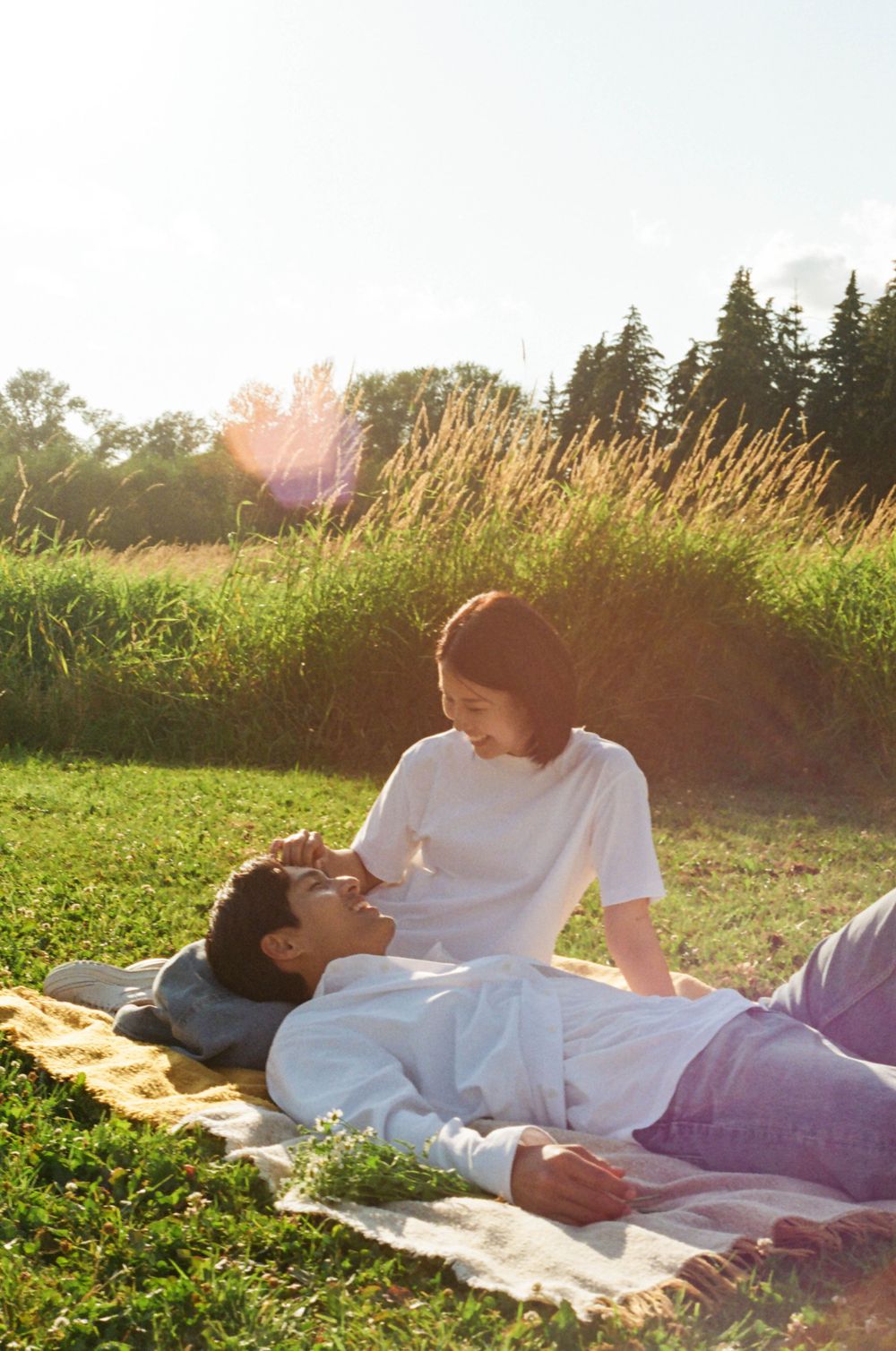 Two people lying on a blanket in a sunny field surrounded by tall grass.