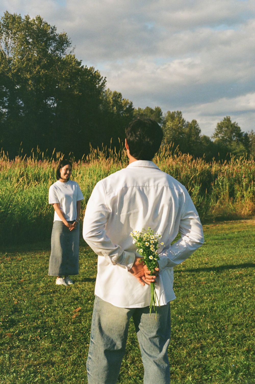 Person holds bouquet of white flowers behind back while another stands in summer meadow.