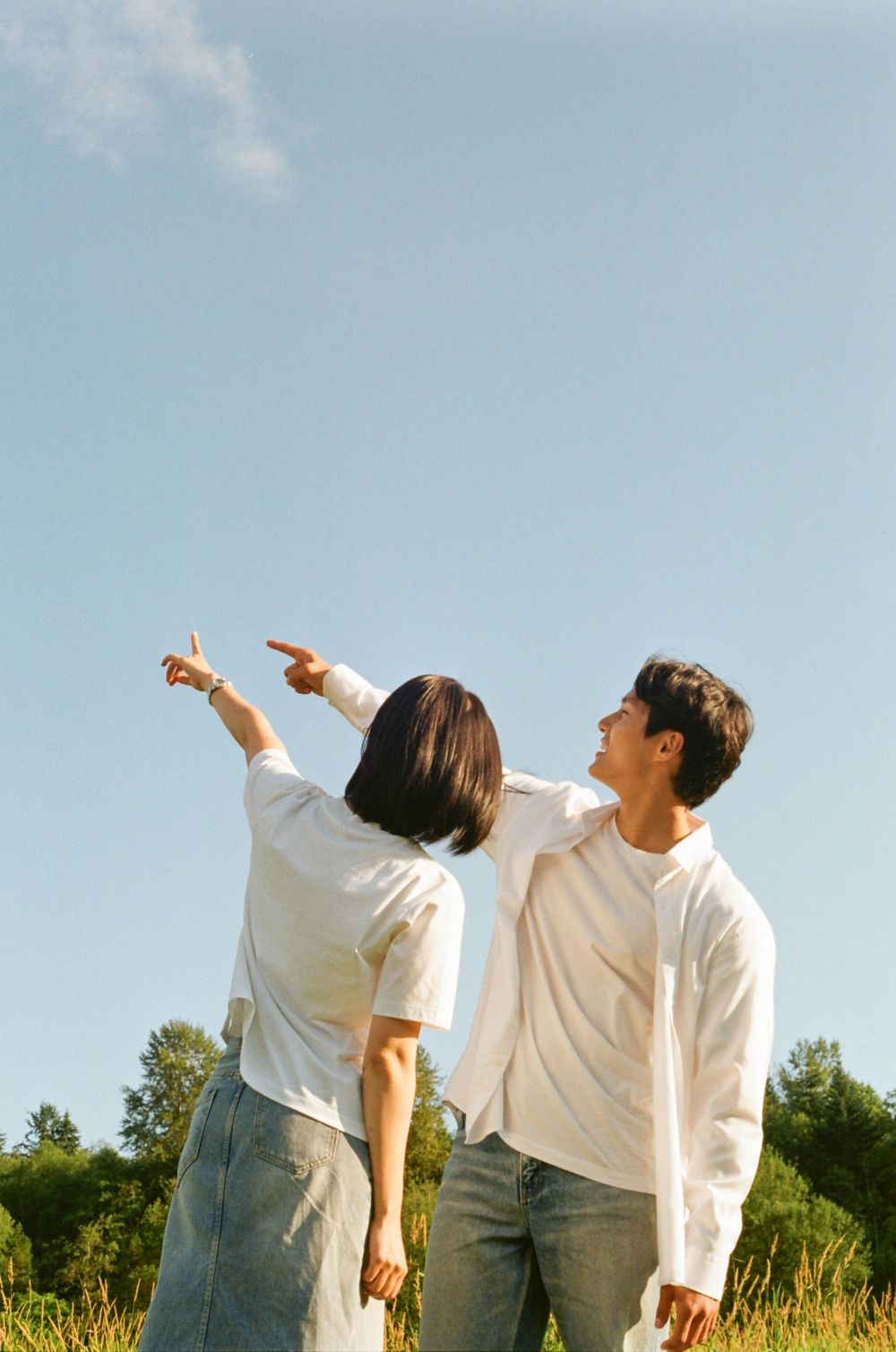 Two people in white shirts and jeans dance playfully in a sunny field with trees in the background.
