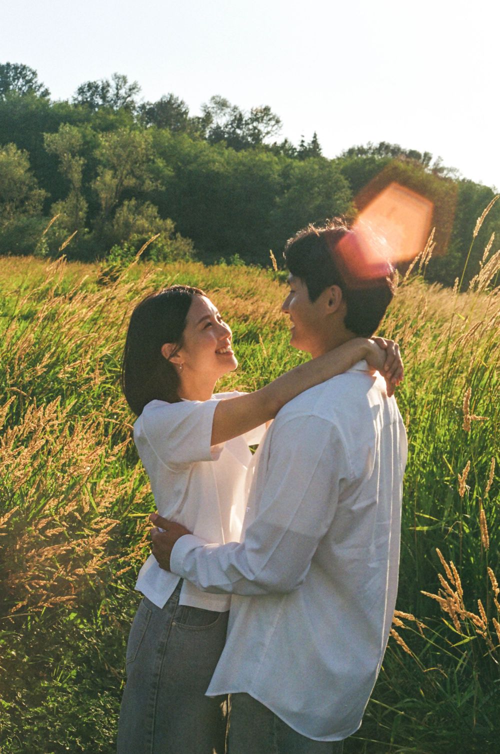 A couple in white clothing embraces playfully in a sunlit field of tall grass during golden hour.