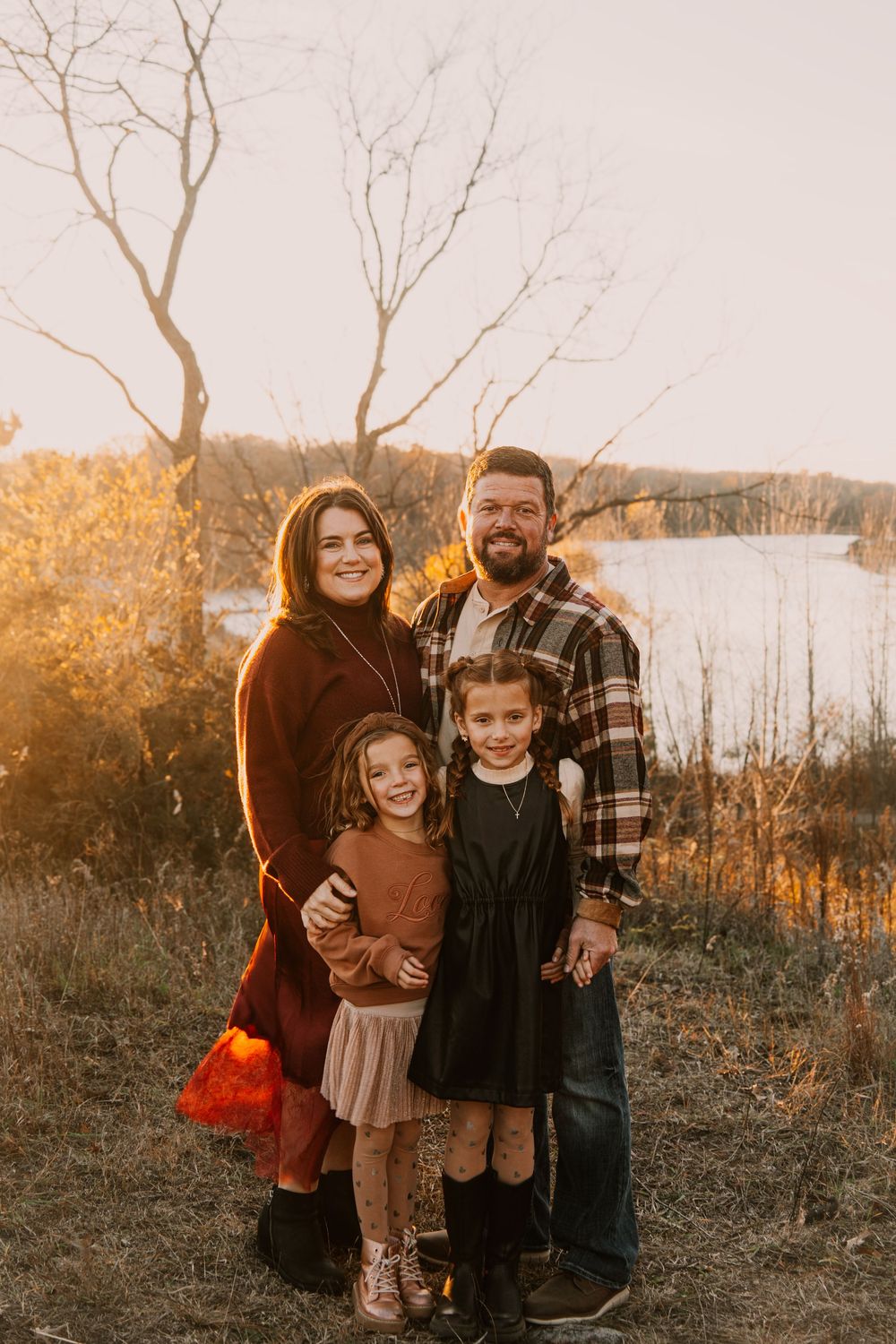 Family portrait at sunset with golden light filtering through bare trees by a lake.