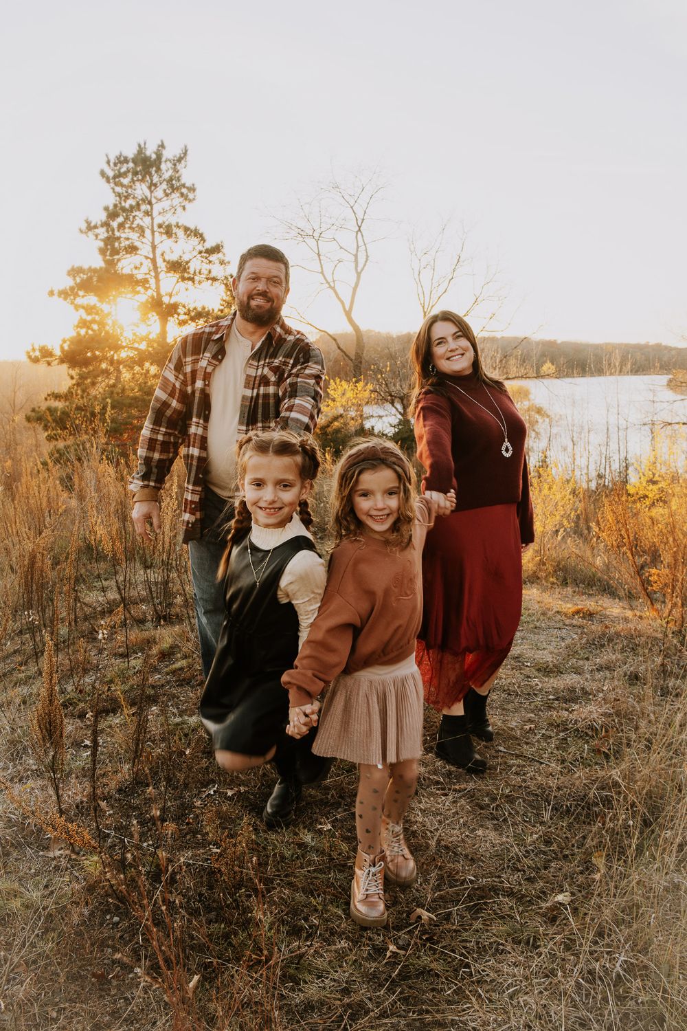 A family in warm earth-toned outfits poses together during golden hour by a lake in autumn.