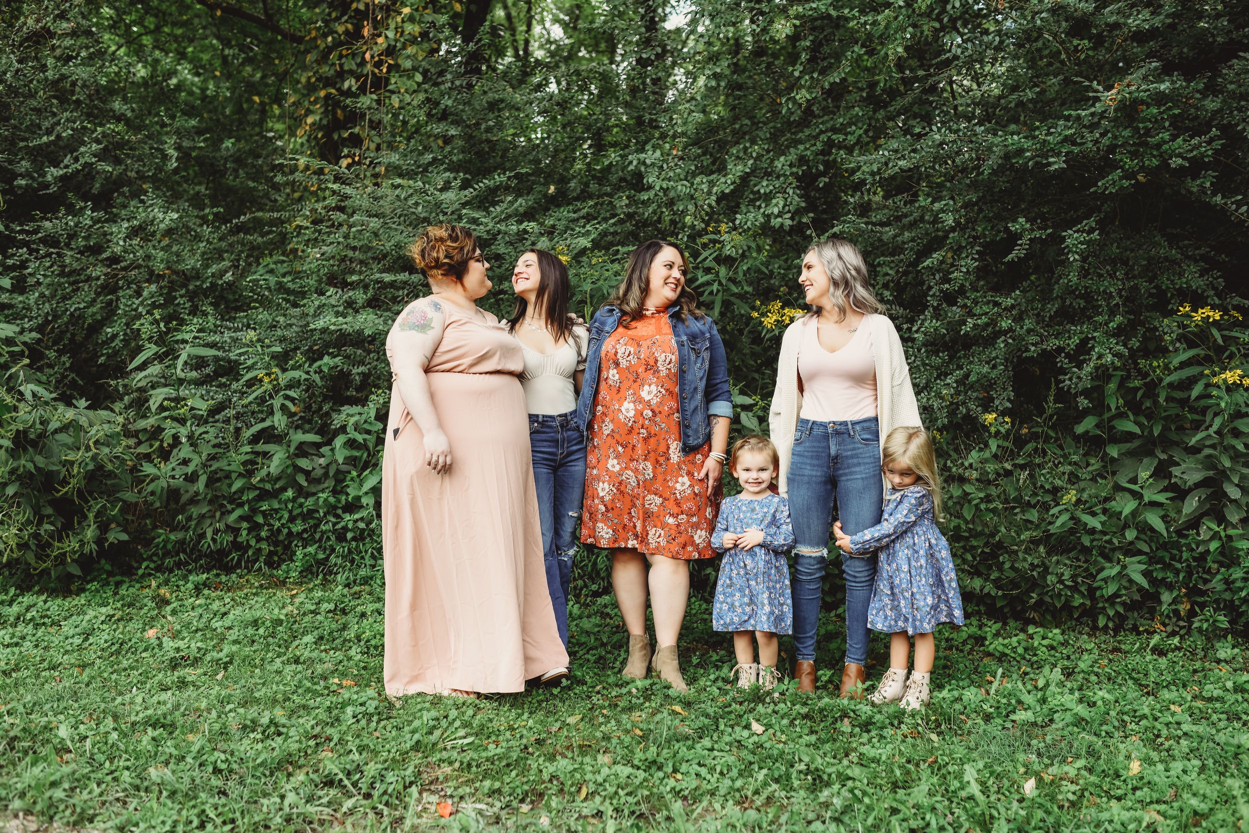 A group poses together outdoors wearing coordinated pastel and floral outfits against green foliage.
