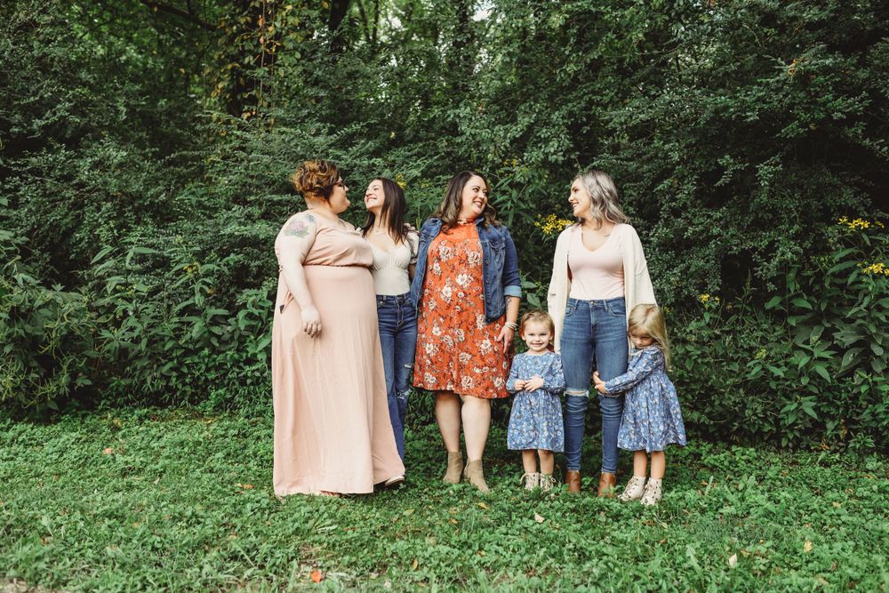 A group poses together outdoors wearing coordinated pastel and floral outfits against green foliage.