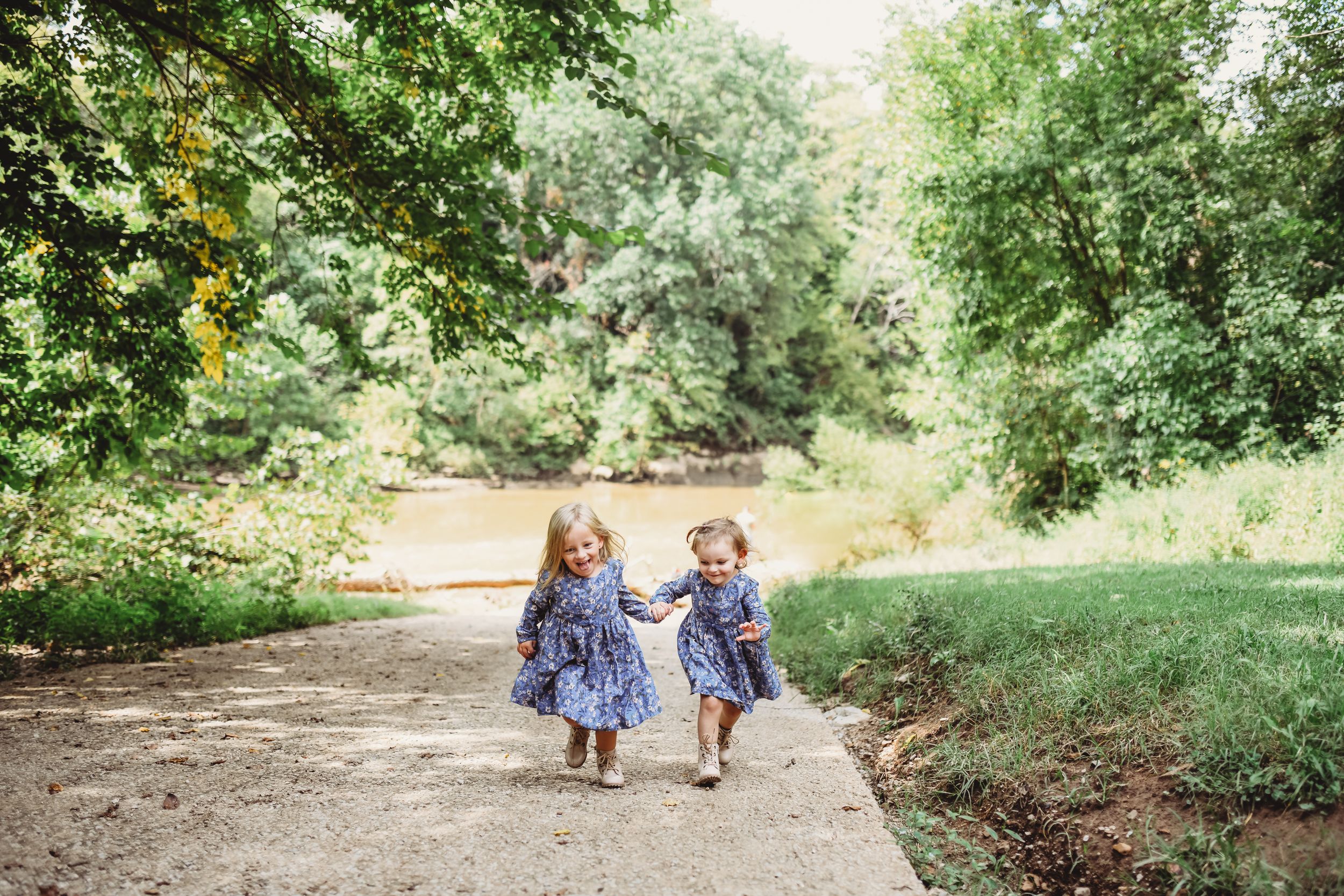 Two children in matching blue dresses run joyfully down a wooded path together.