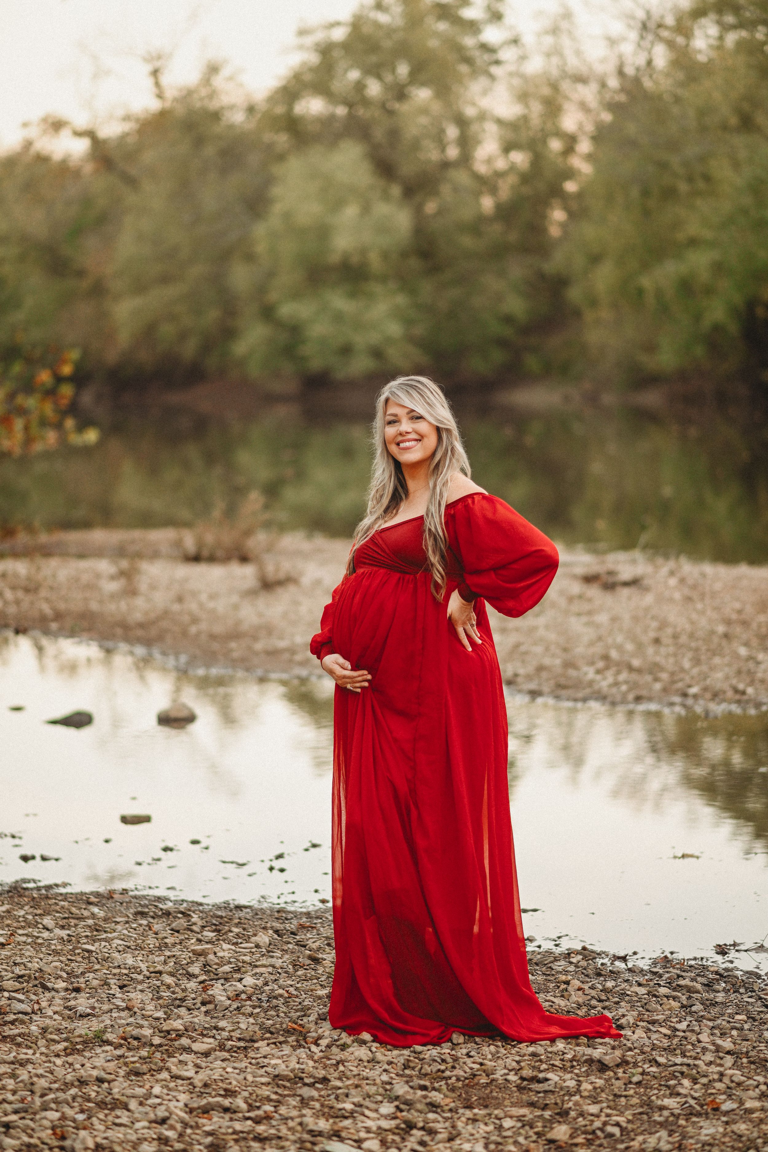 Woman in flowing red dress poses gracefully by water's edge at sunset.