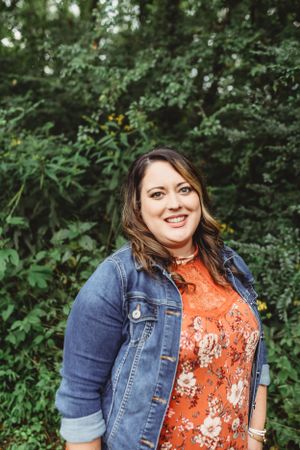 A woman in a denim jacket and floral orange top smiles outdoors against lush greenery.