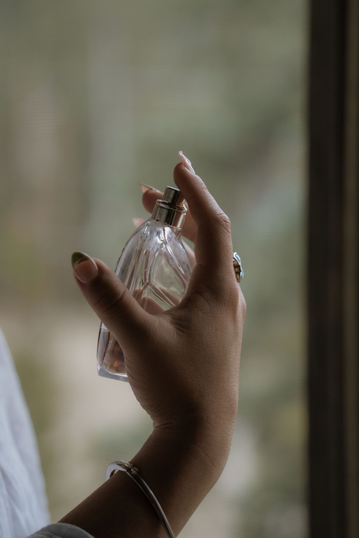 Hand holds a glass perfume bottle against a blurred green background in natural lighting.