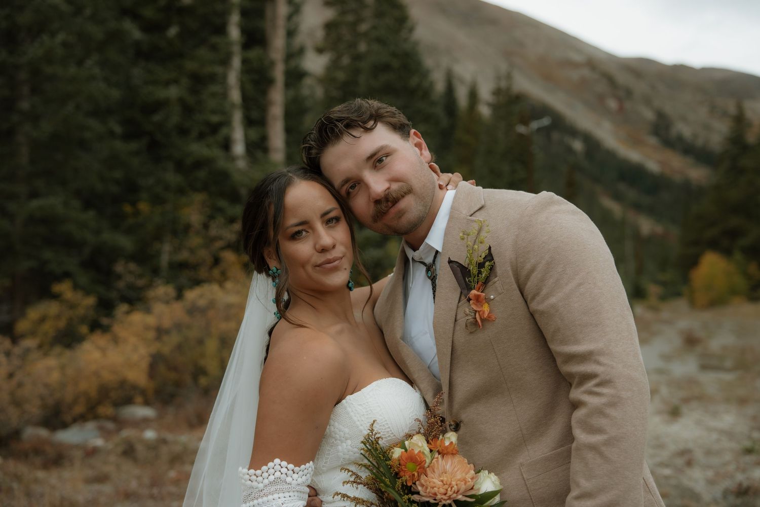 A couple in wedding attire embrace in front of a mountainous autumn landscape with evergreen trees.