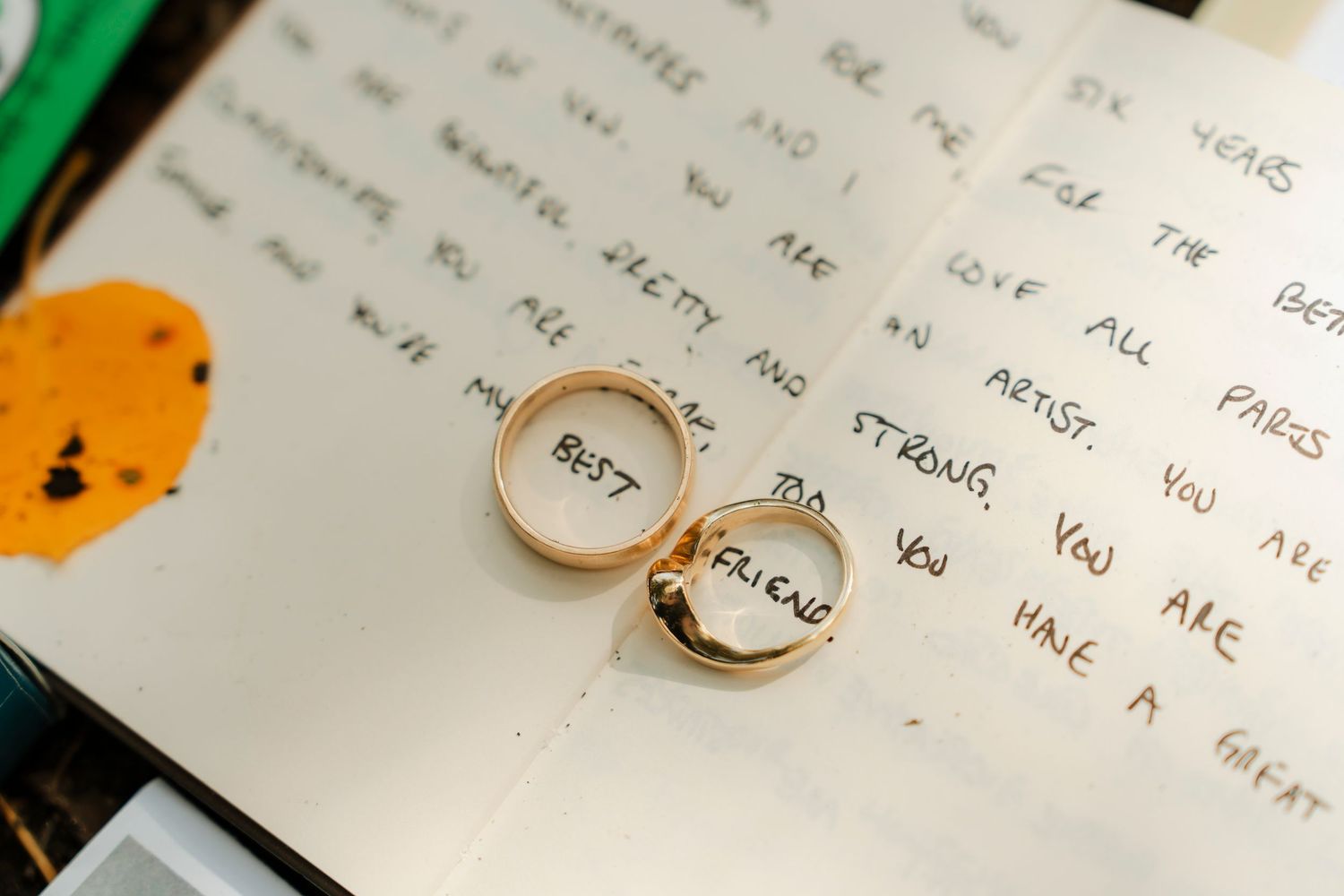 Two gold wedding rings with text engraved on them resting on handwritten notes or letters.