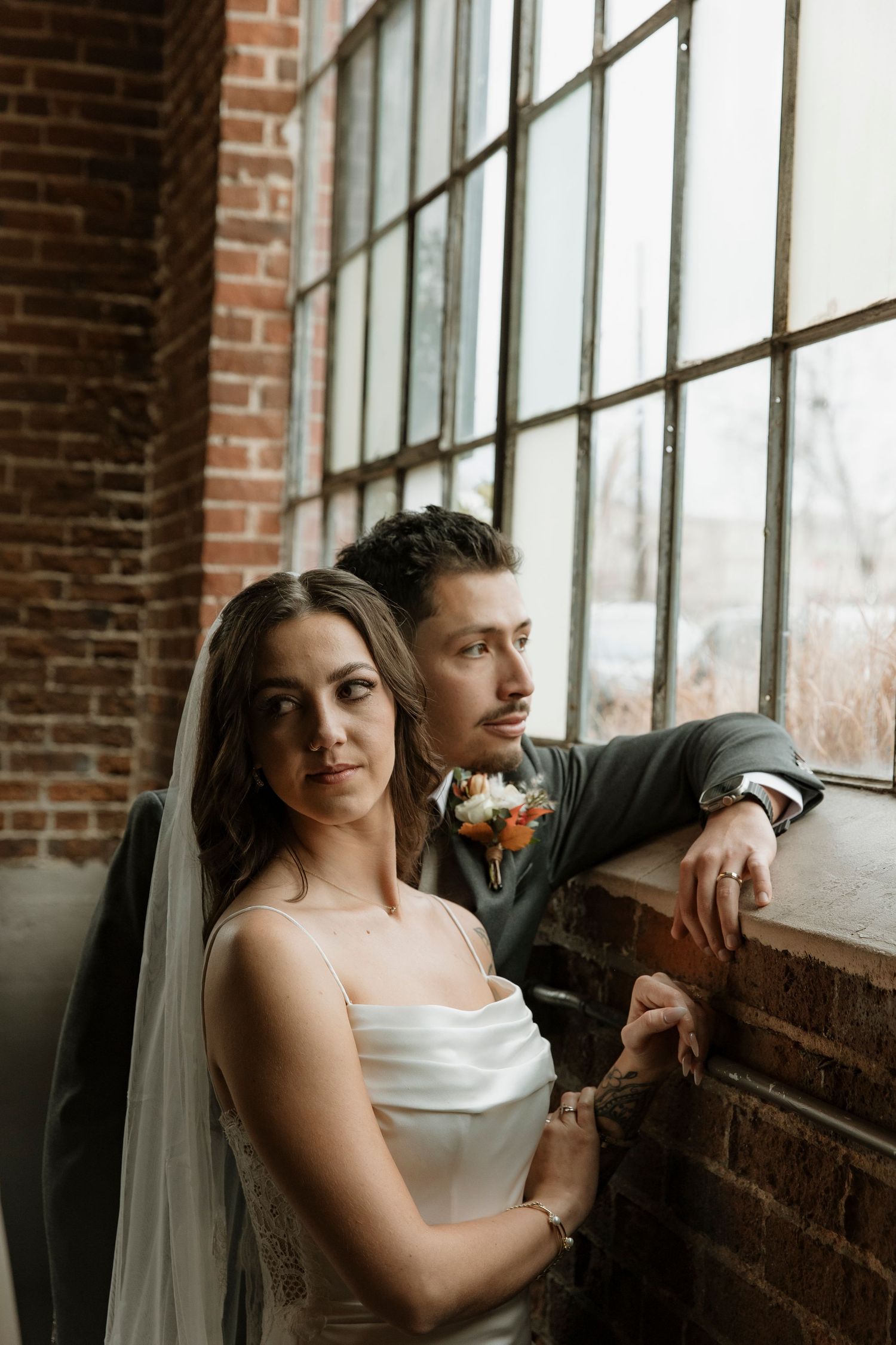 A bride and groom pose together near a large industrial window in a rustic brick venue.