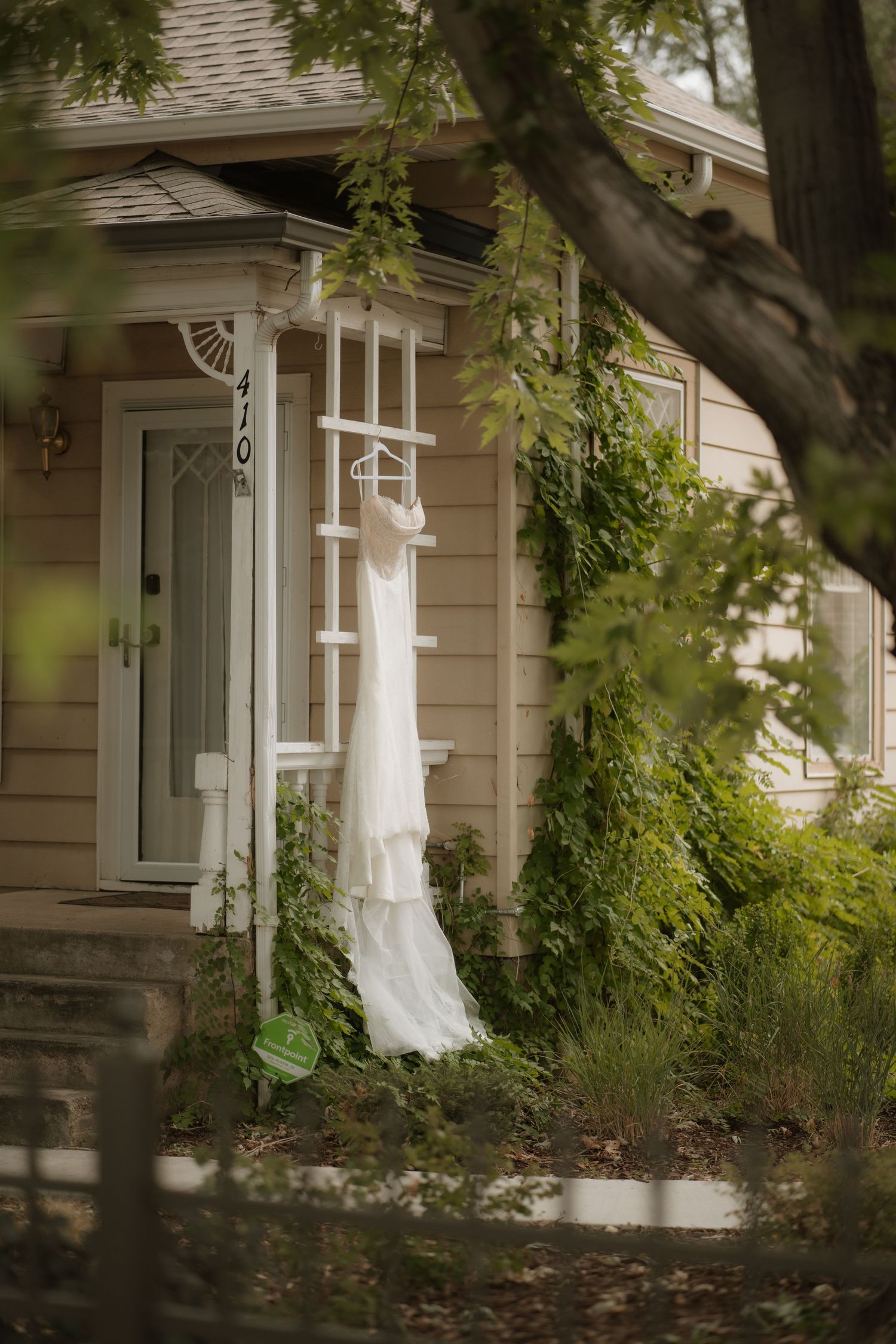 A white wedding dress hangs elegantly outside a charming craftsman-style house with green foliage in the foreground.