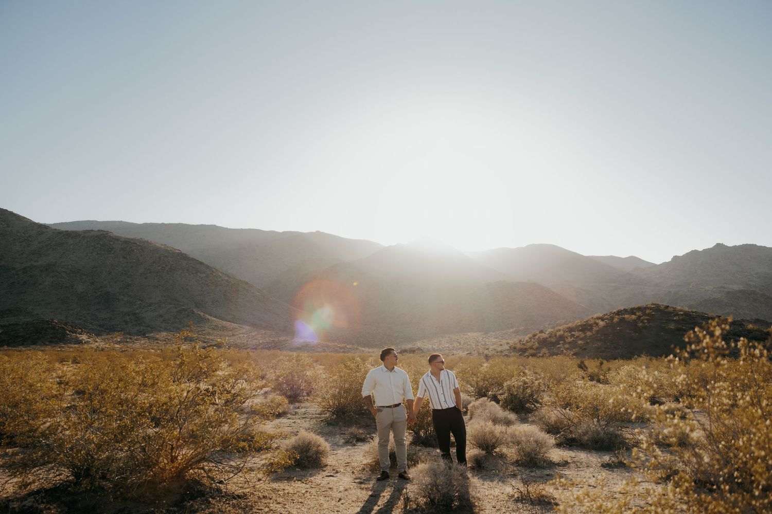 Couple hiking through desert terrain with mountains and sunlight in background.