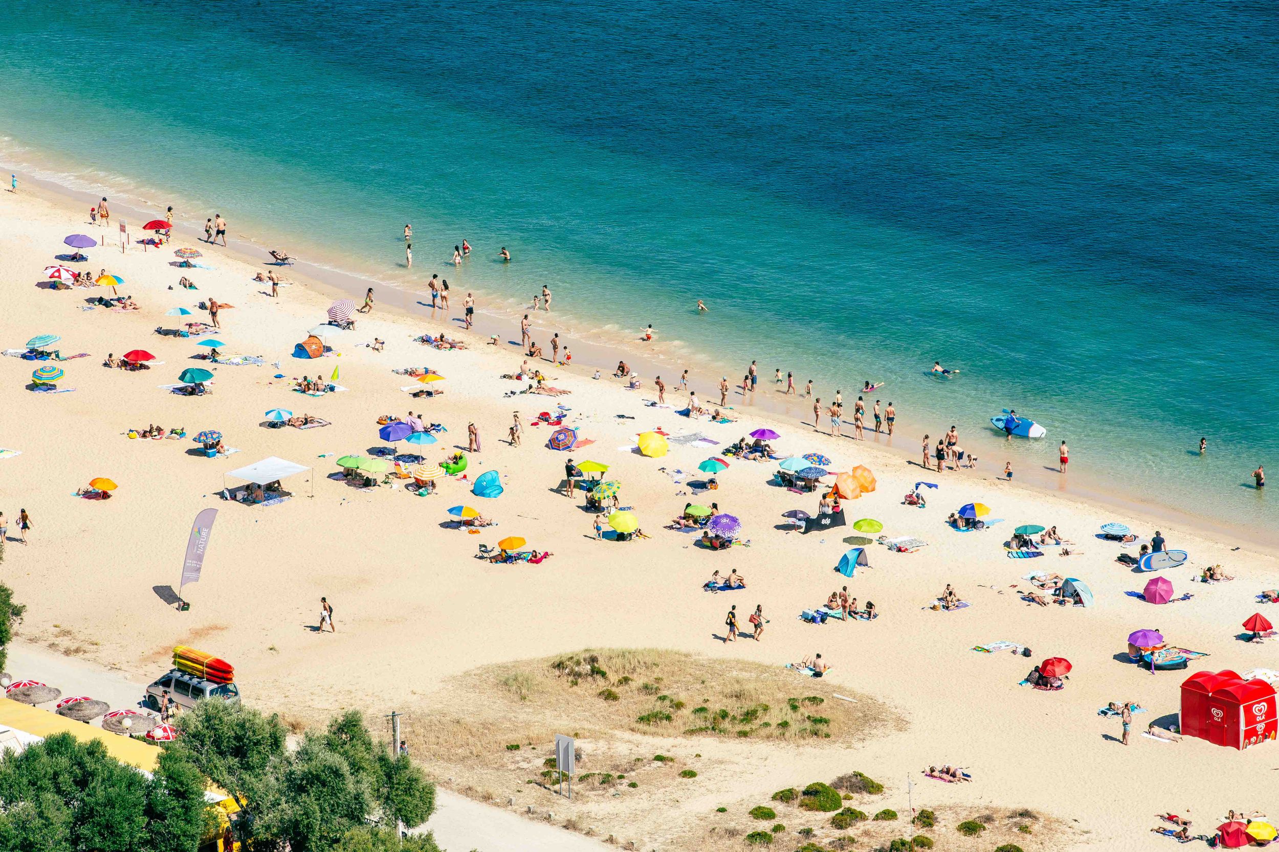 Colorful beach umbrellas dot the golden sand alongside turquoise waters viewed from above.