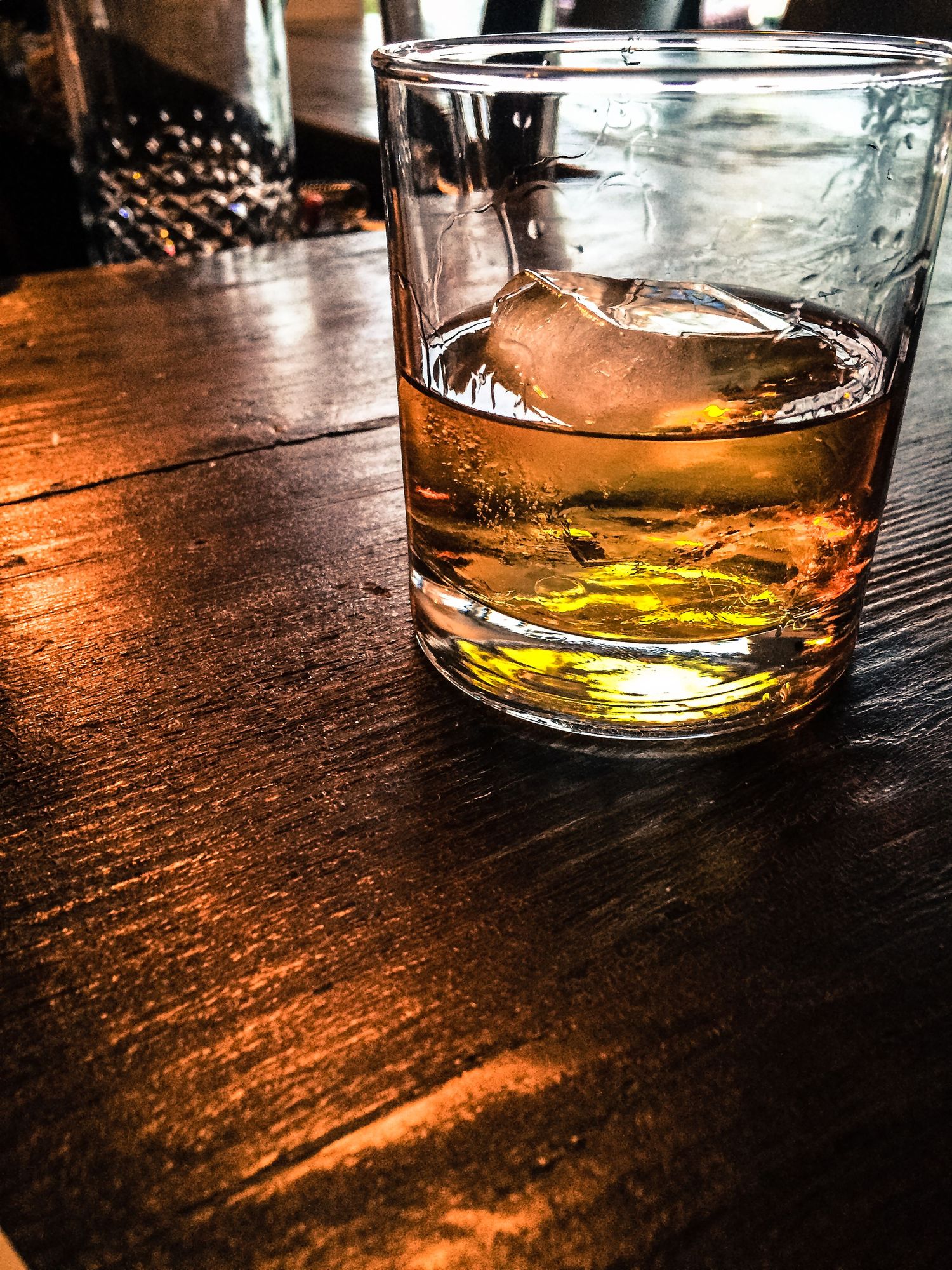 A glass of amber-colored whiskey with ice on a dark wooden table.