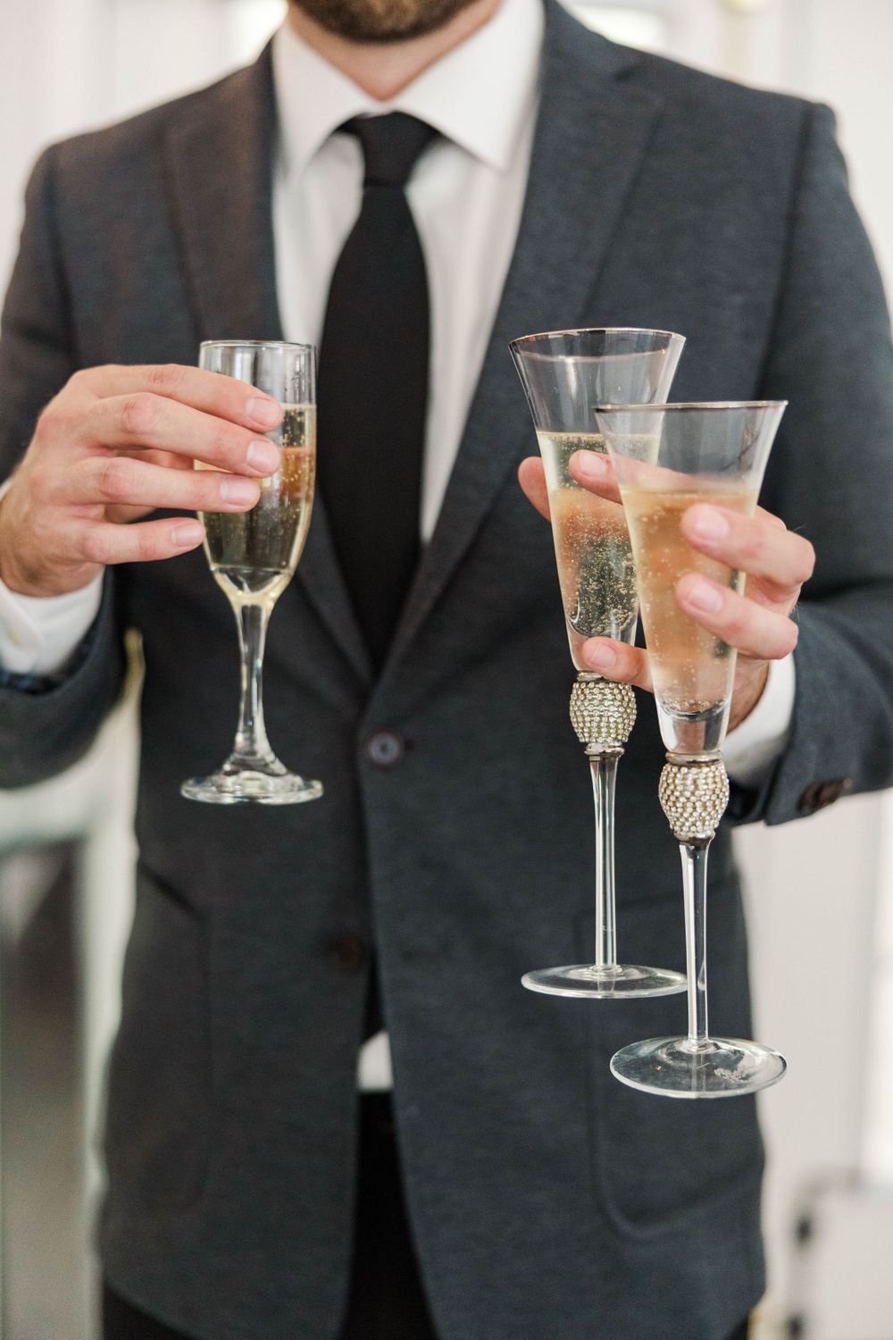 Groom in gray suit holding three champagne flutes at wedding reception.