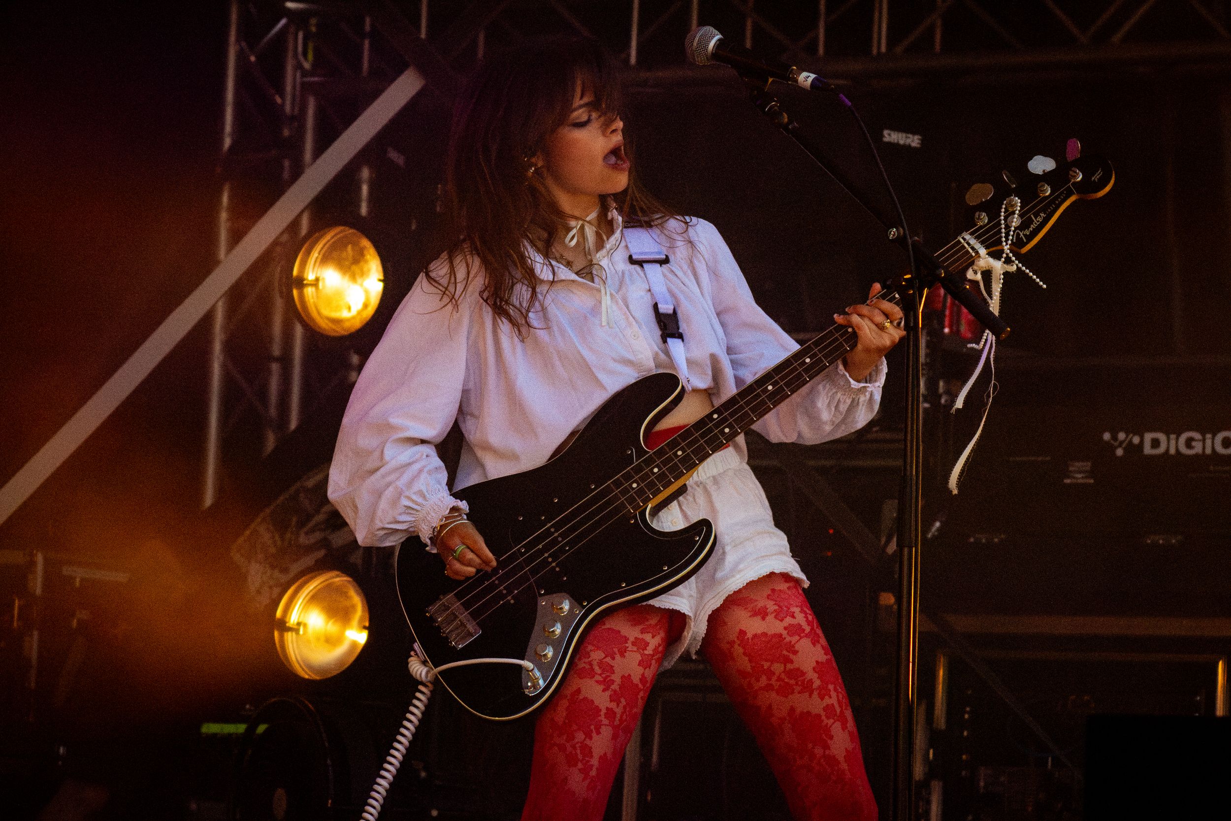 Performer plays bass guitar on stage wearing white shirt and red pants with amber stage lights.