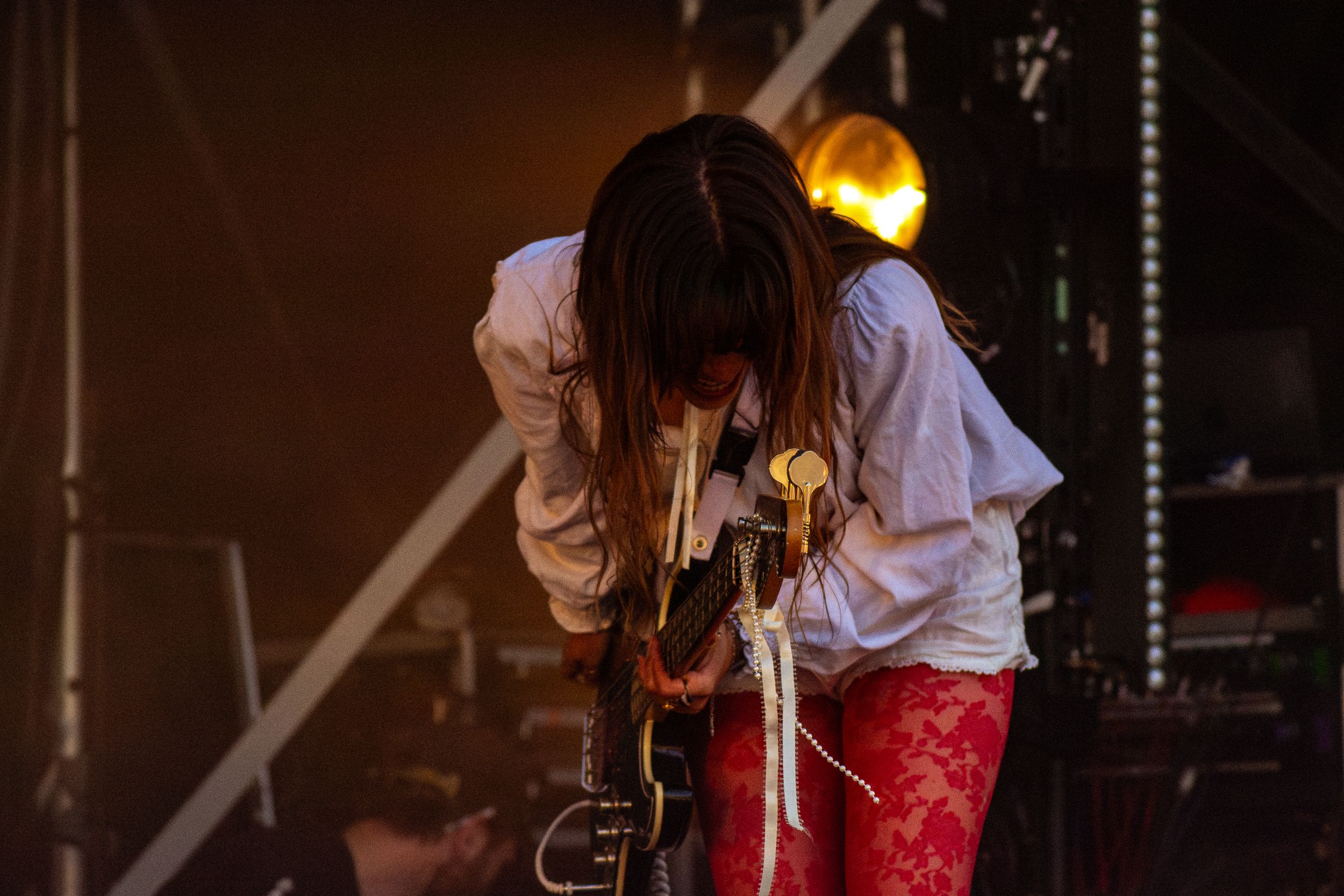 Performer bends forward while playing guitar on stage in white shirt and red pants.