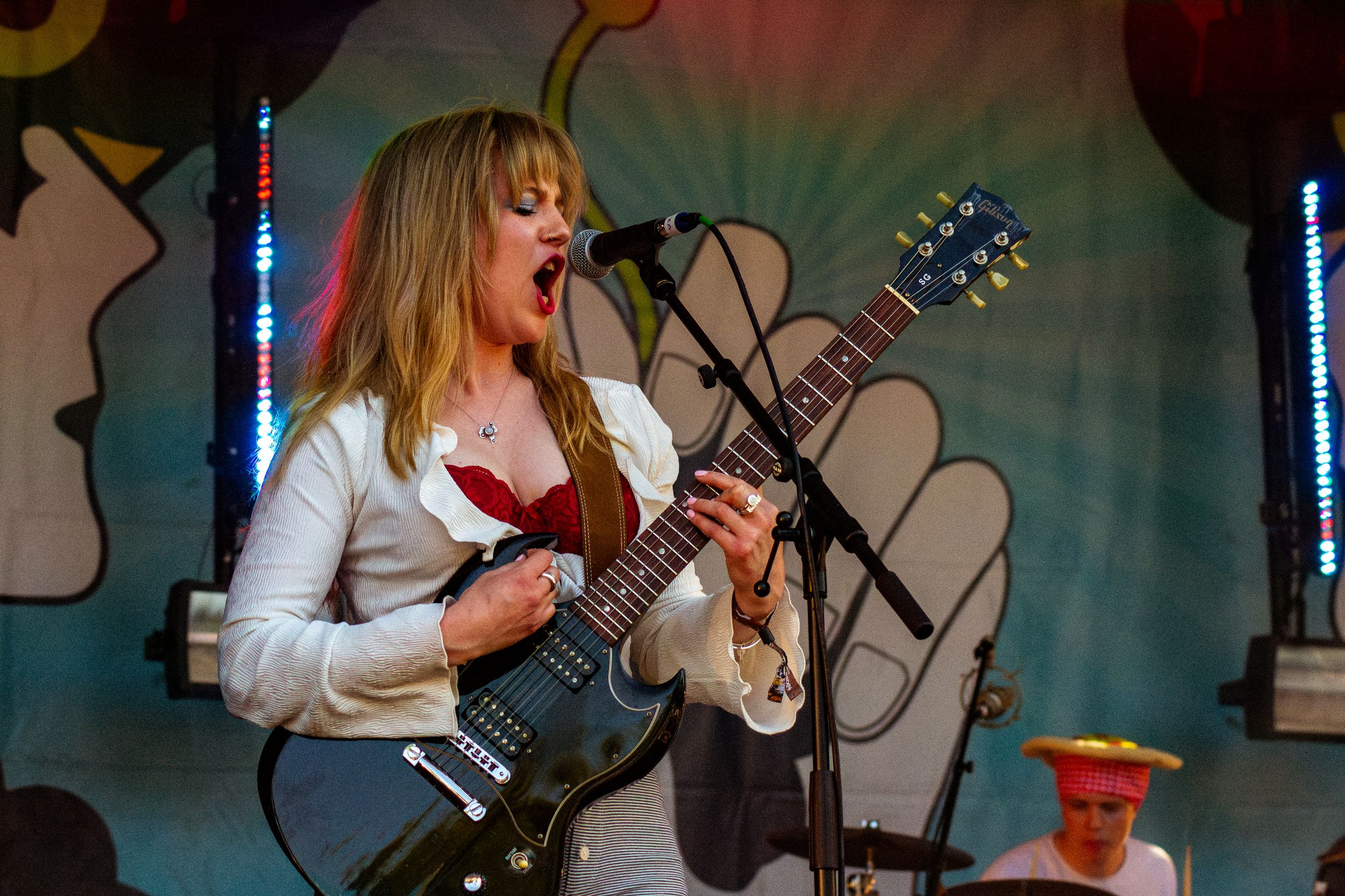 Performer plays electric guitar on stage with decorative flower backdrop and colorful lighting.