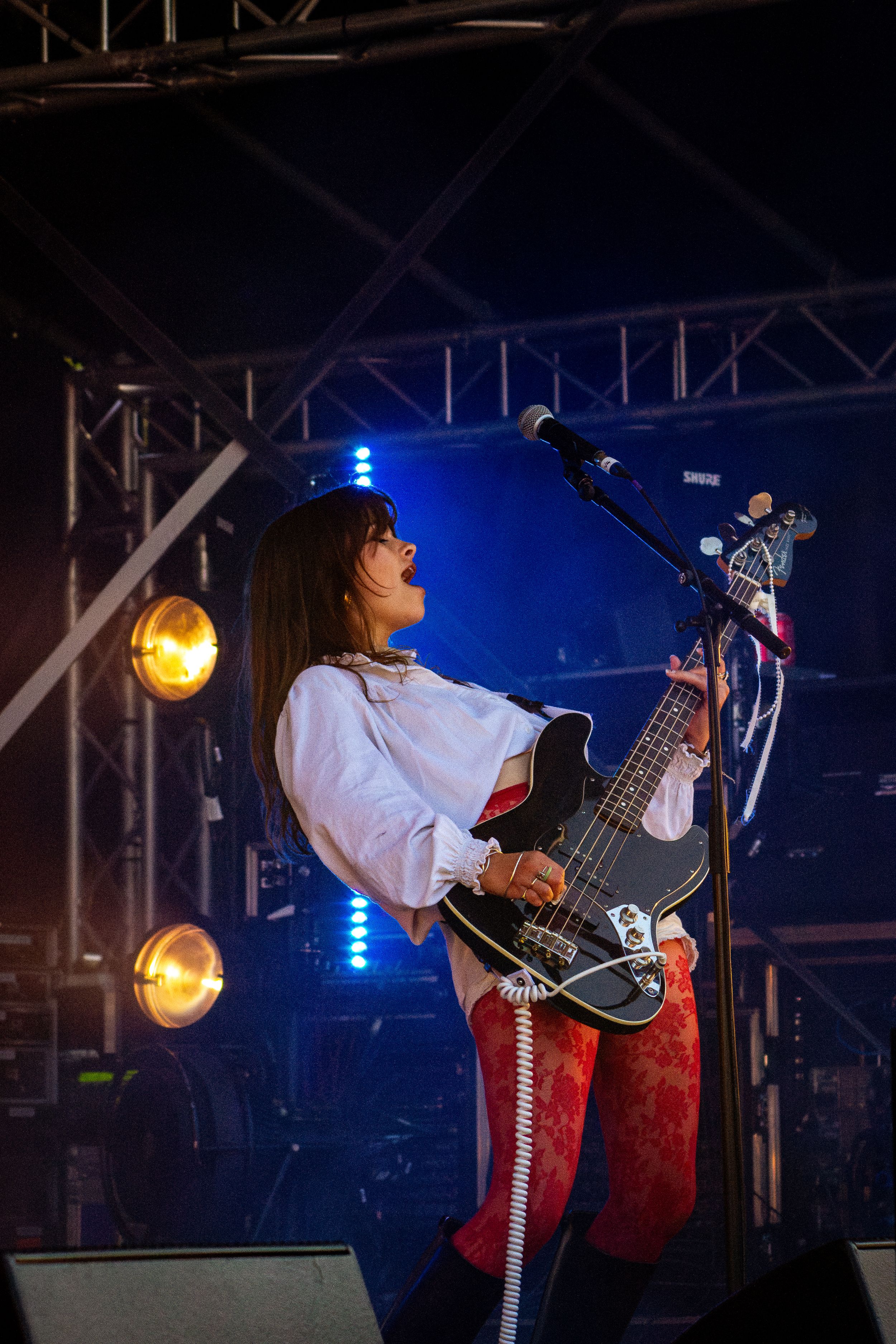 Musician plays bass guitar on stage wearing white shirt and red pants with dramatic lighting.