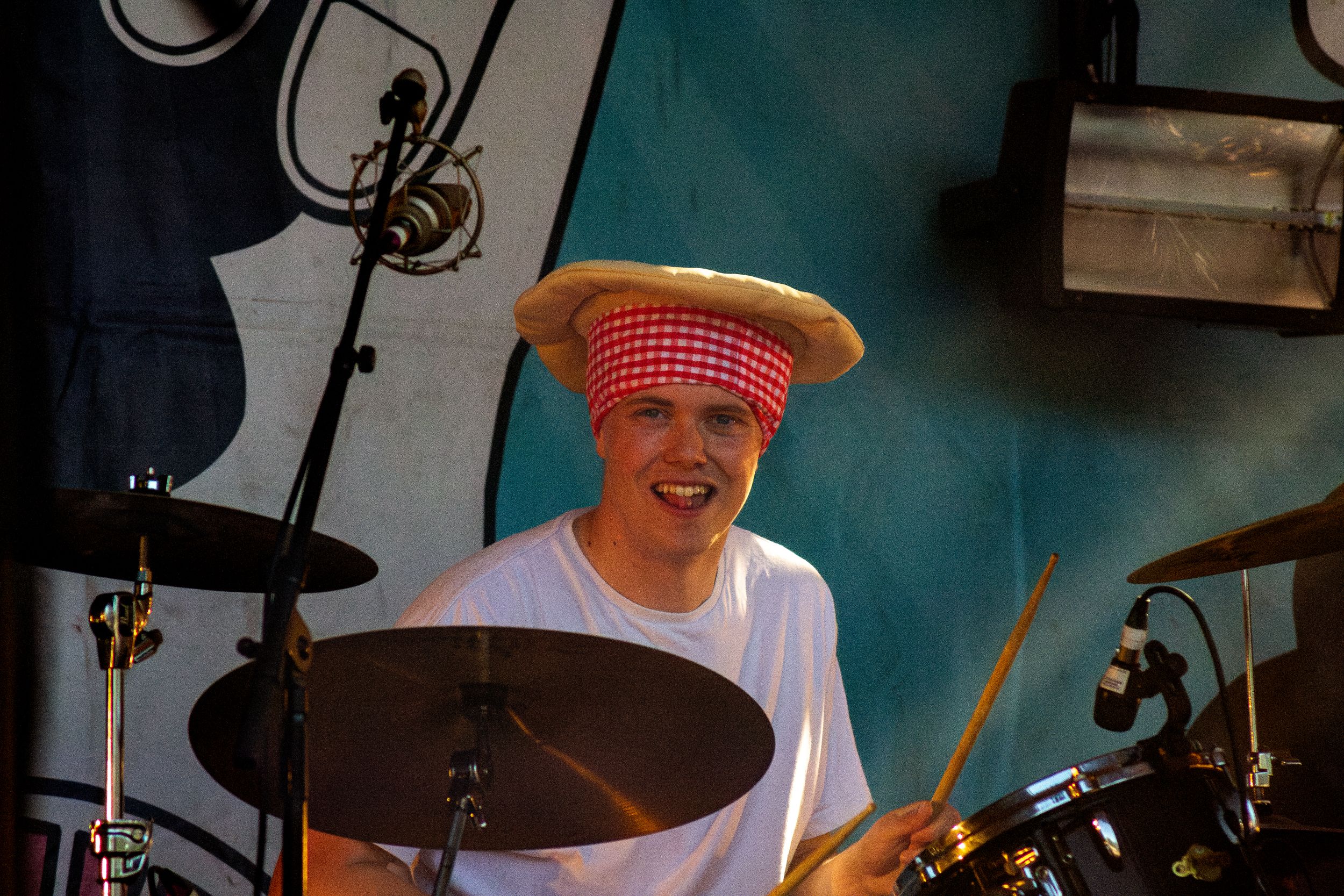 Drummer performs wearing red hat and white shirt behind drum kit on stage.