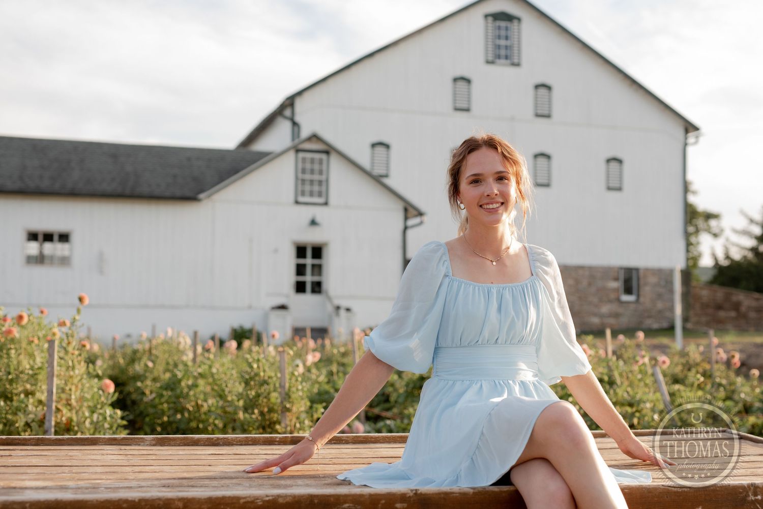 A flowing light blue dress creates contrast against a rustic white barn and wooden bench setting.