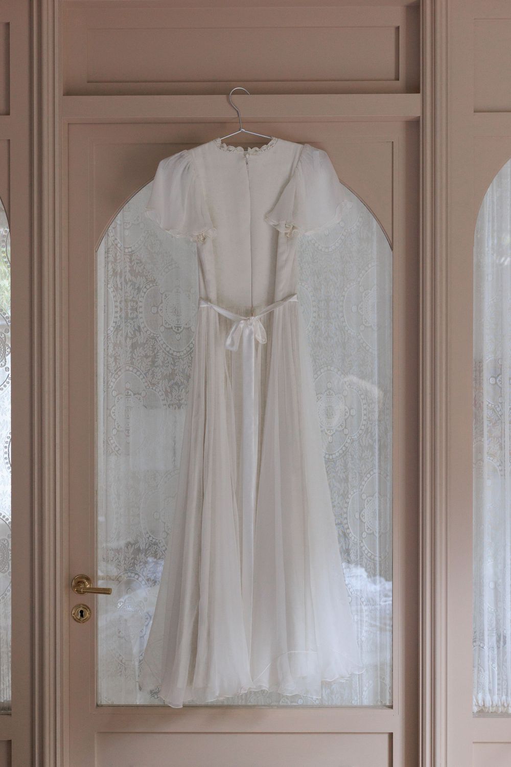 A delicate white wedding dress with lace details hangs on a hanger against a beige paneled door.