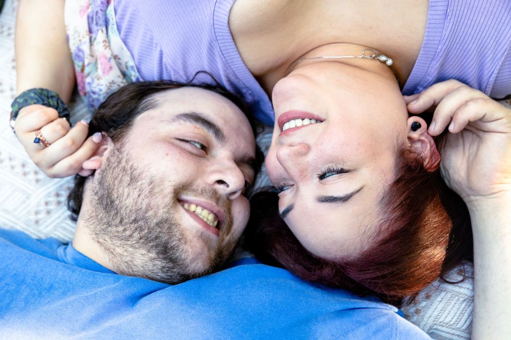 A couple lies together on a blanket outdoors sharing a playful moment.