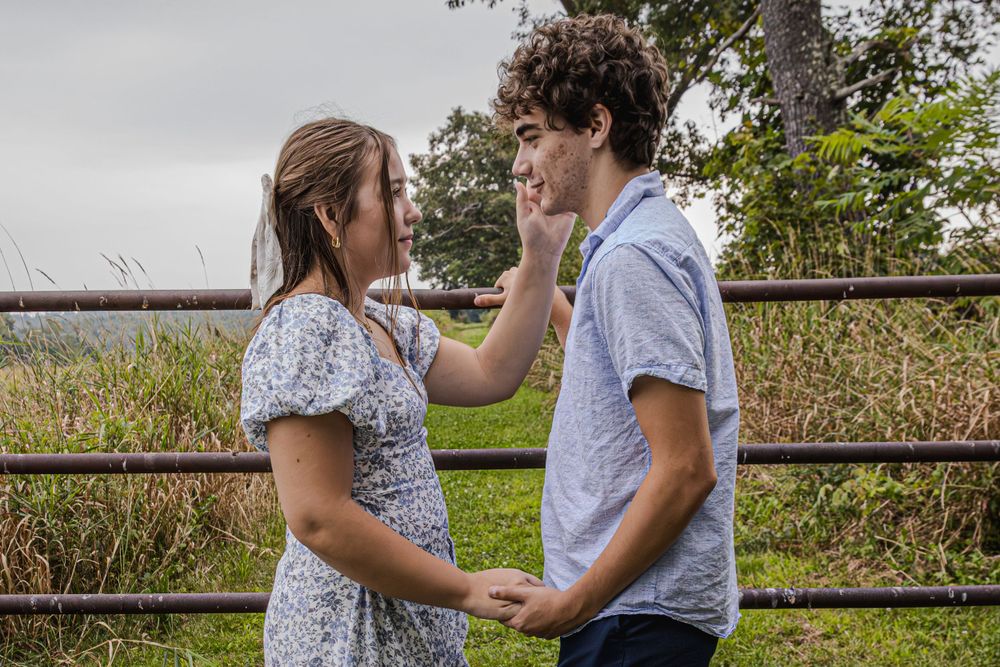 A couple stands by a wooden fence on a hilltop overlooking scenic countryside views.