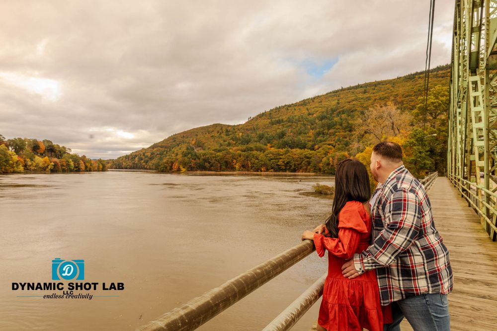 A couple shares a romantic moment on a bridge overlooking a river with autumn foliage in the background.