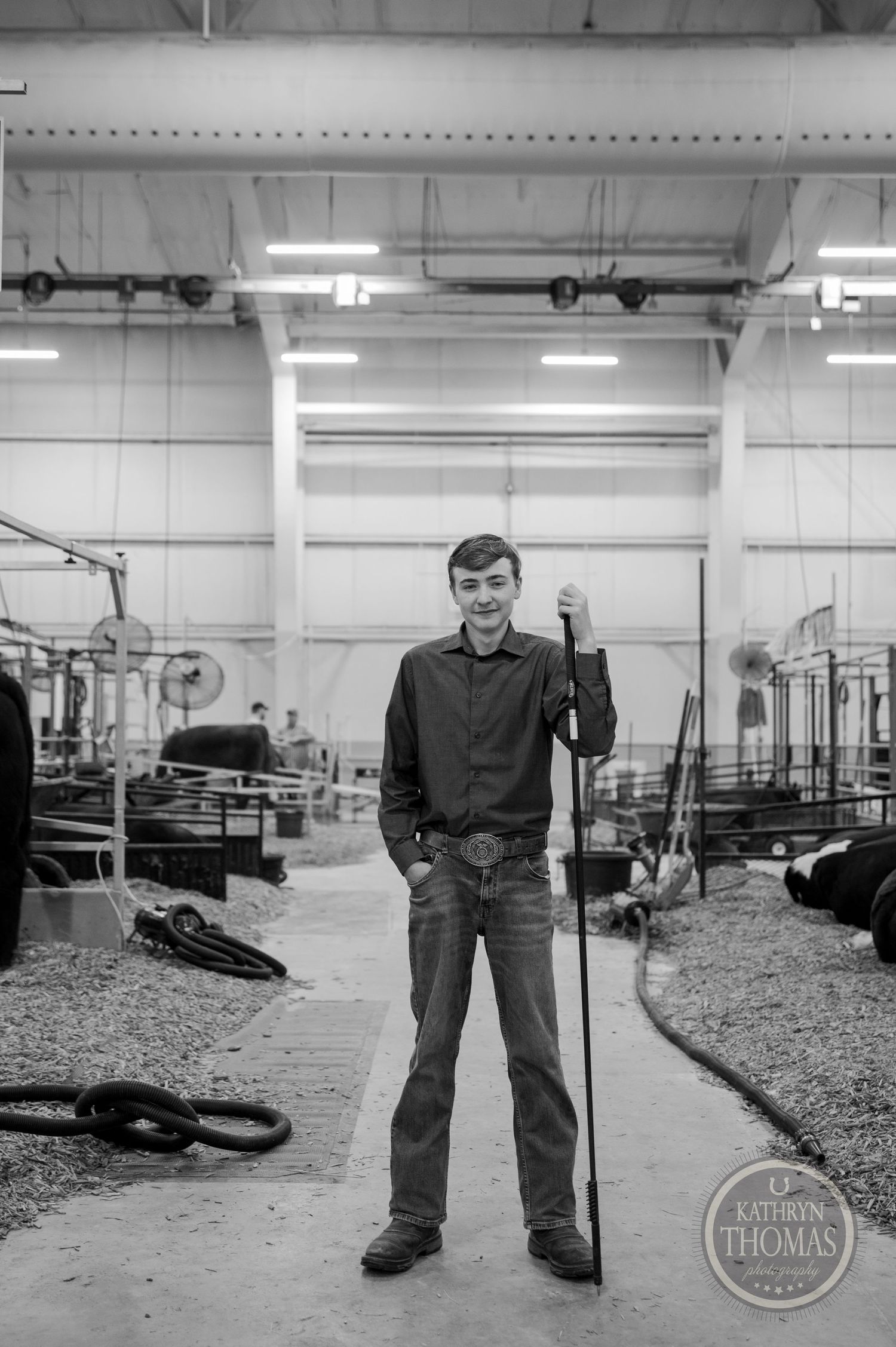 A person stands with a show staff in an indoor livestock facility with equipment and supplies visible.