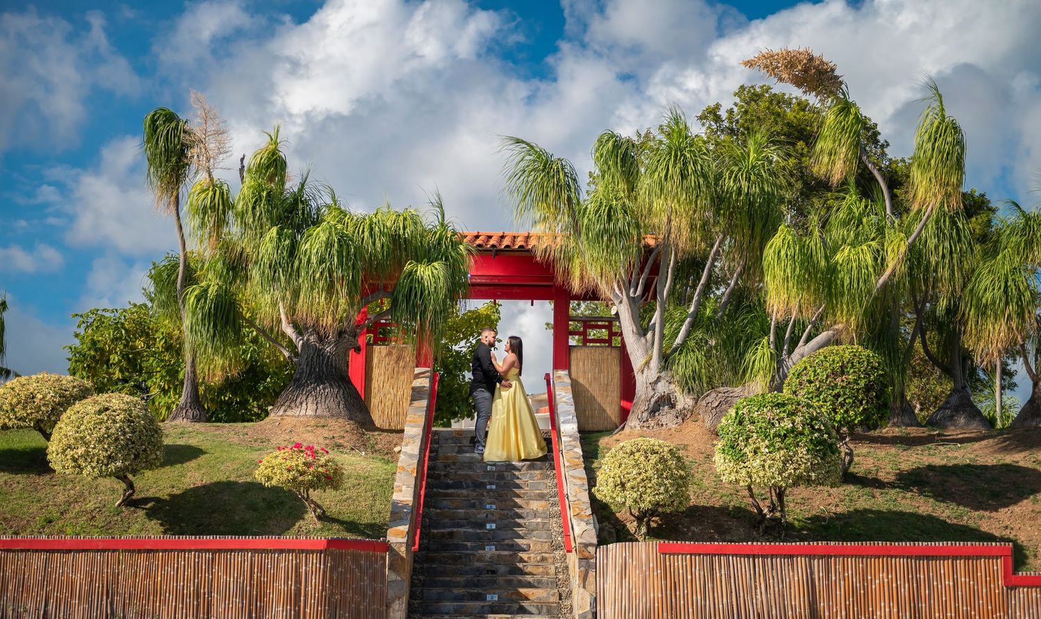 Red oriental gateway with stone steps surrounded by palm trees and tropical landscaping against blue sky.