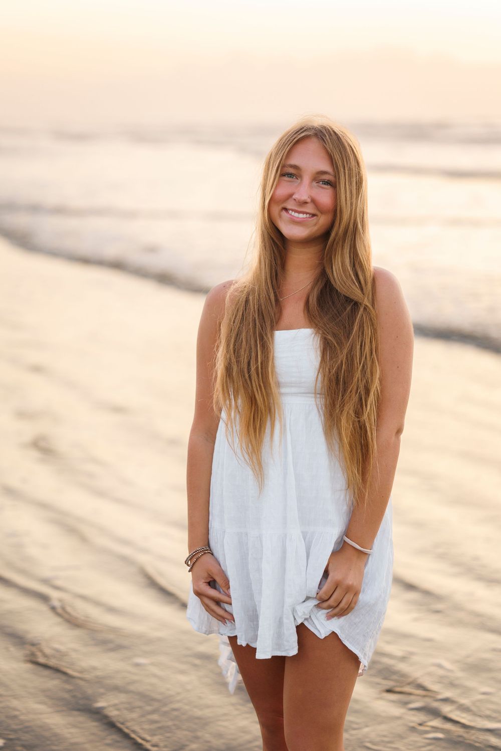 Person in flowy white summer dress standing on beach at golden hour sunset with ocean waves in background.