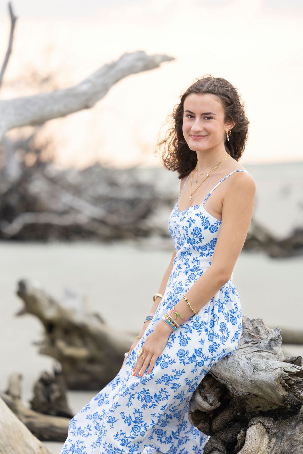 A woman in a blue and white floral sundress poses gracefully on driftwood at the beach during sunset.