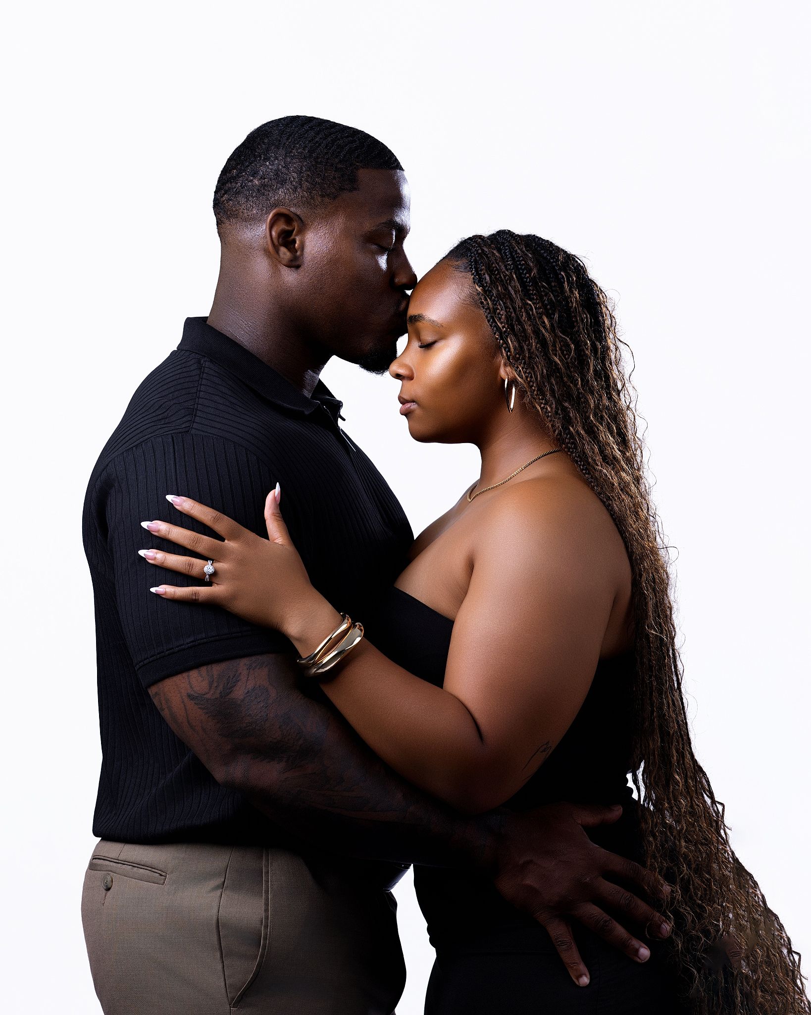 A couple in black outfits sharing an intimate moment, nearly kissing, against a white background.