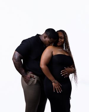 A couple in matching black outfits share an intimate embrace against a white background in a maternity photo shoot.