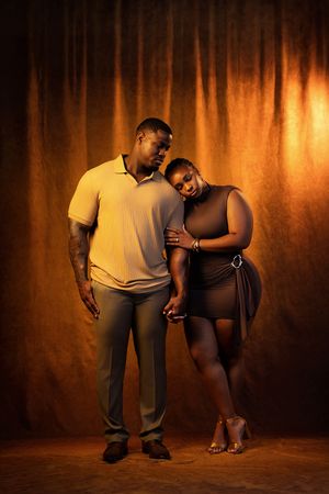 A couple poses intimately in a studio, the woman leaning on the man, both dressed in earth-toned outfits against a warm backdrop.