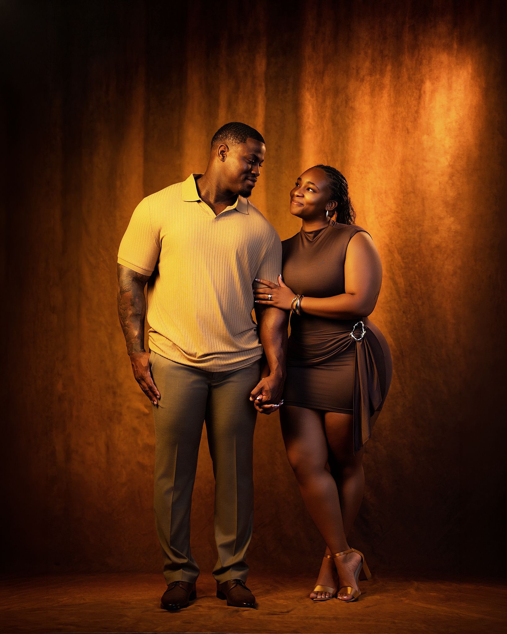 Couple posing together in warm golden studio light, man in yellow polo and woman in brown mini dress holding hands.