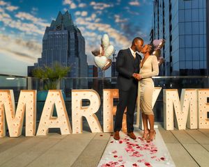 Couple kissing in front of lit MARRY ME sign on rooftop after engagement proposal.