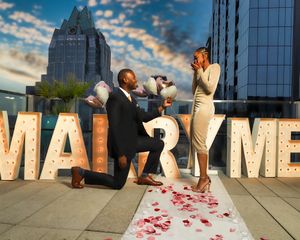 Man on one knee proposing to woman beside large MARRY ME marquee letters at sunset.