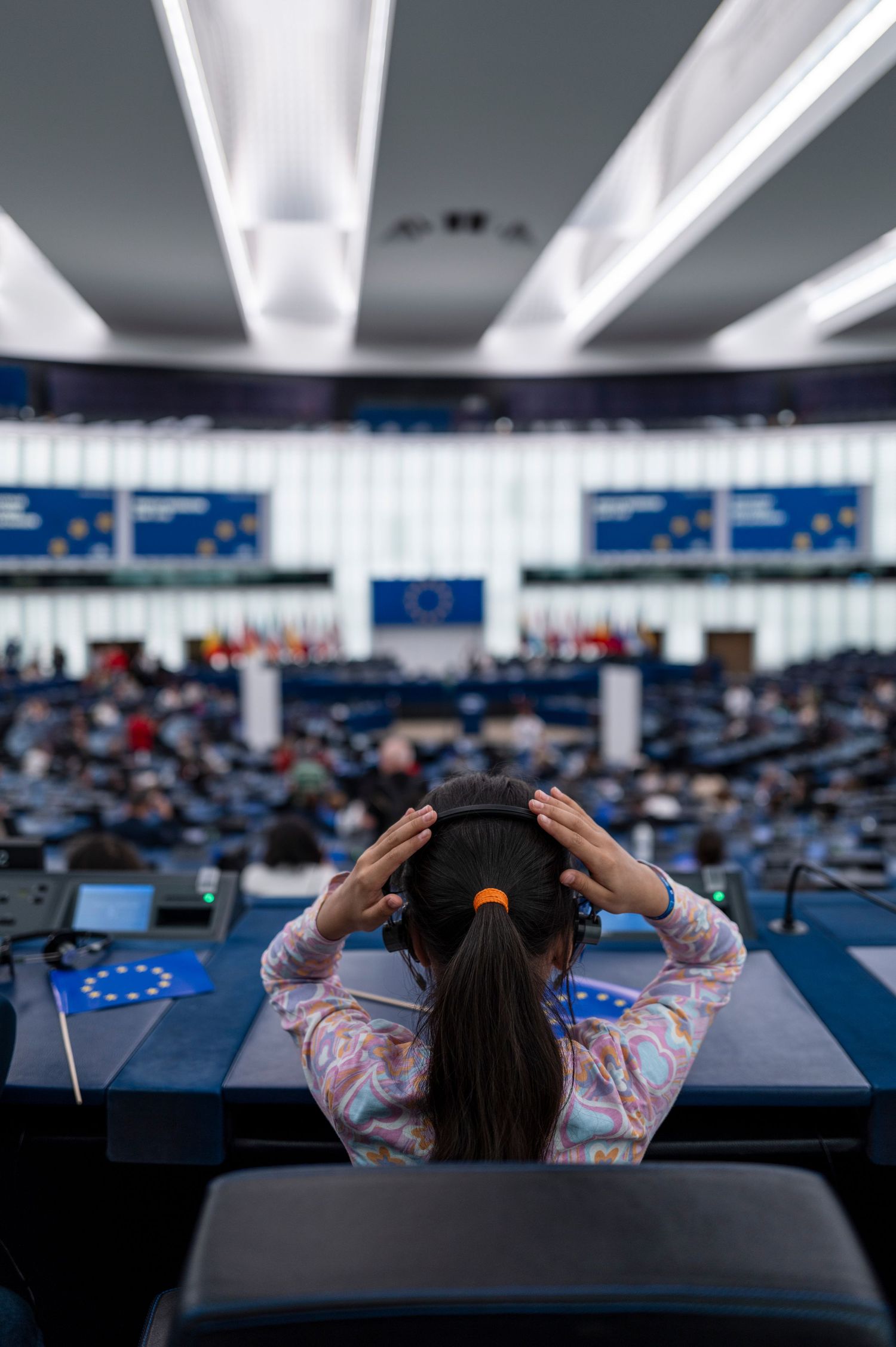 A person sitting in the European Parliament chamber with illuminated ceiling lights and EU flags visible in the background.