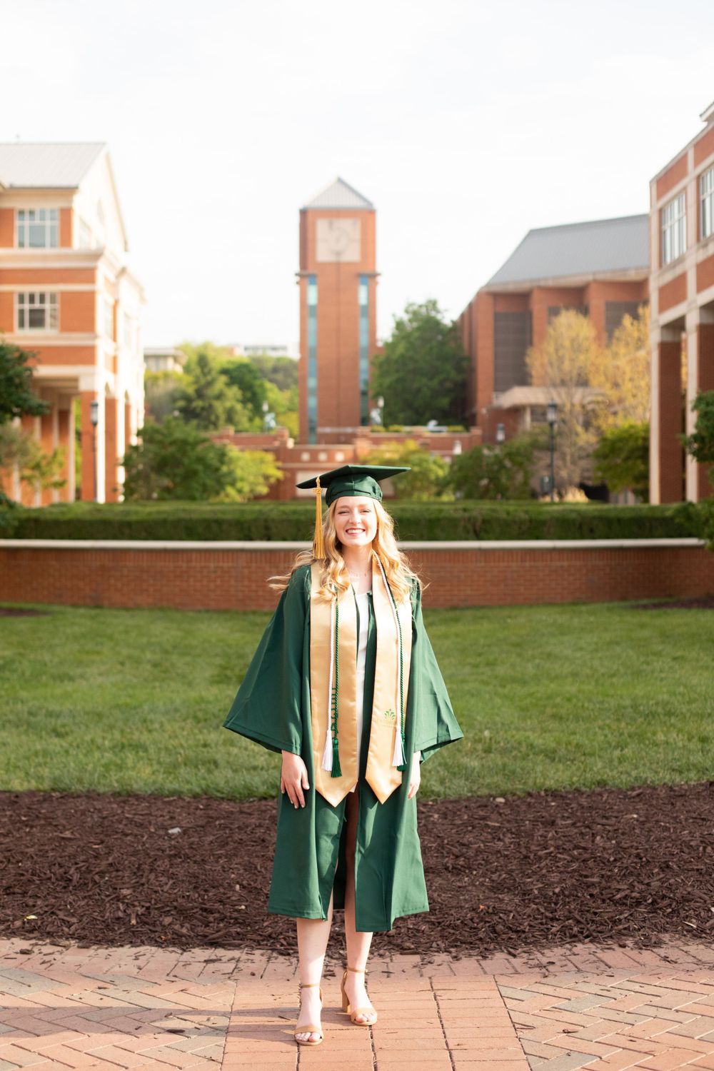 Graduate in green regalia poses on brick pathway with university buildings in background.