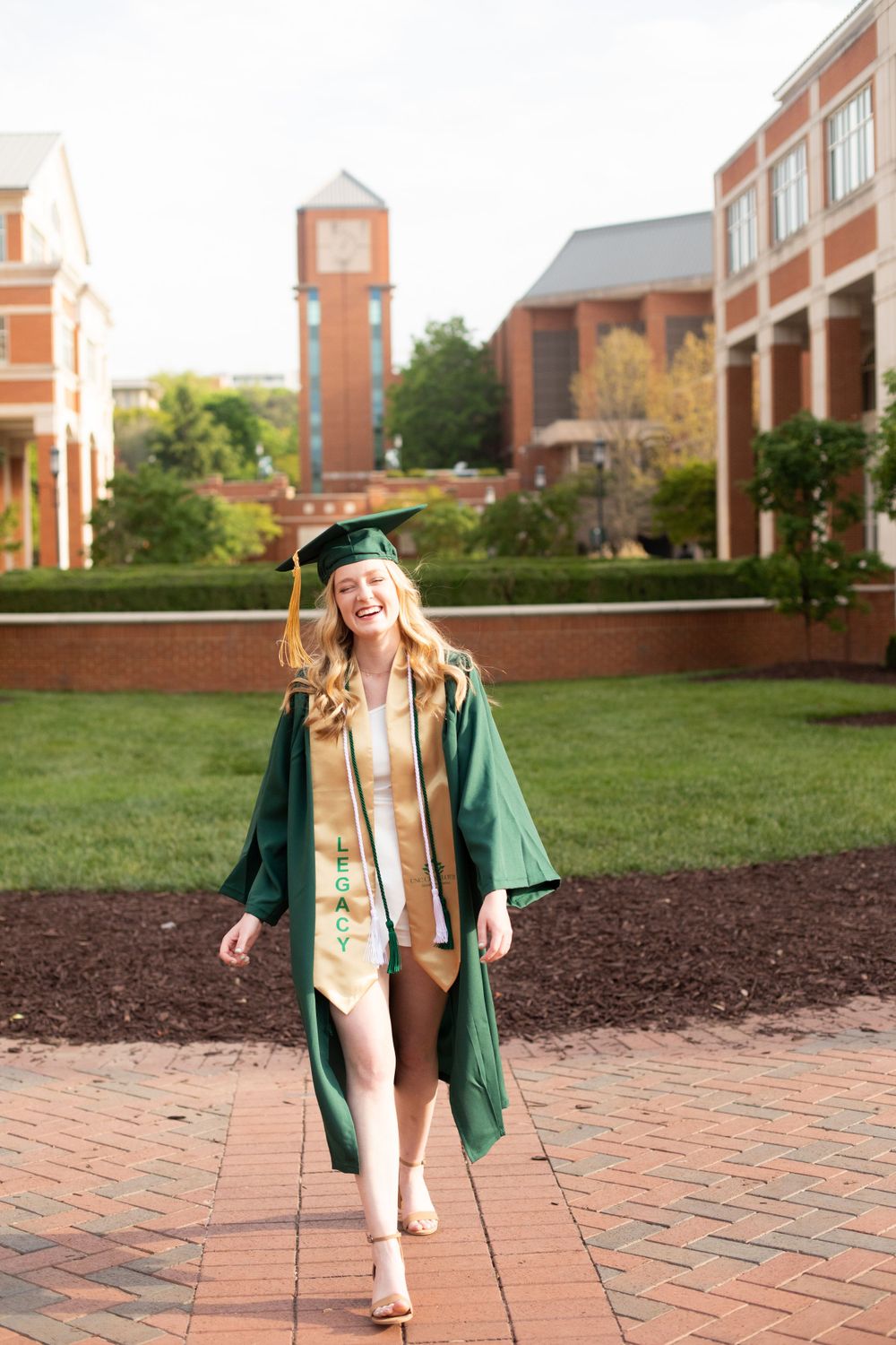 Graduate in green cap and gown walks joyfully across brick courtyard with campus buildings behind.