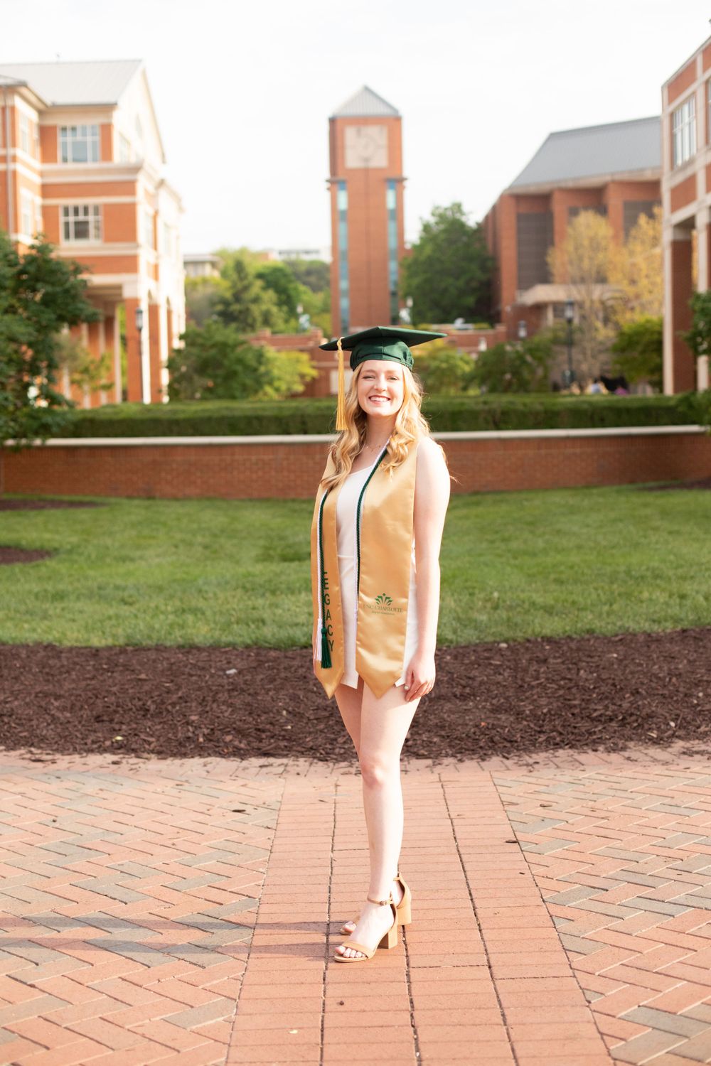 Graduate in green regalia stands confidently on brick pathway with academic buildings in background.