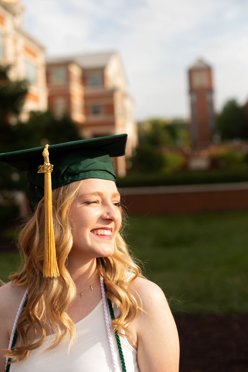 Graduate in green cap with gold tassel smiles brightly during outdoor graduation photoshoot.
